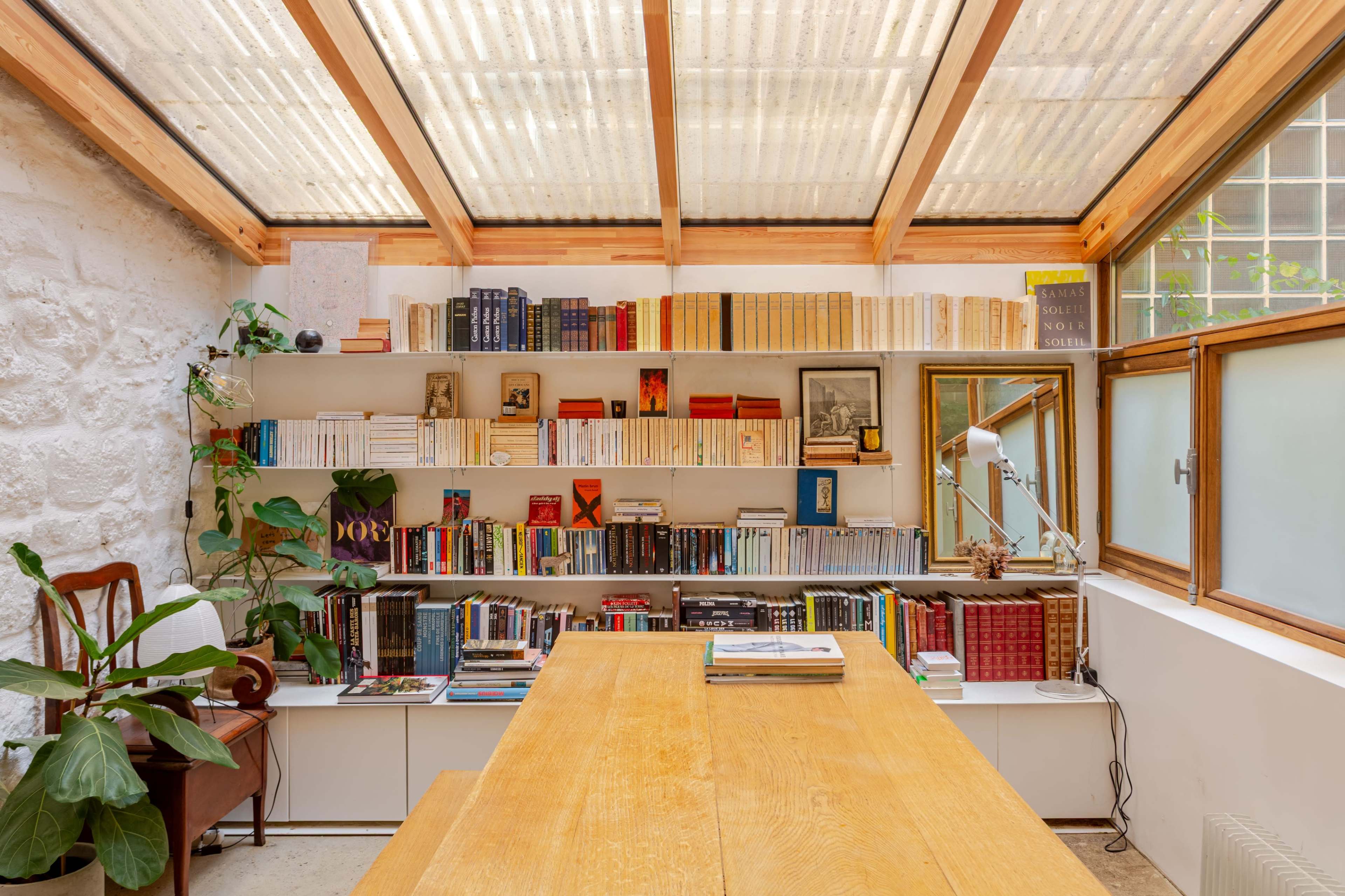 A bright room features a wooden table surrounded by shelves filled with books and a green plant, all under a glass roof.