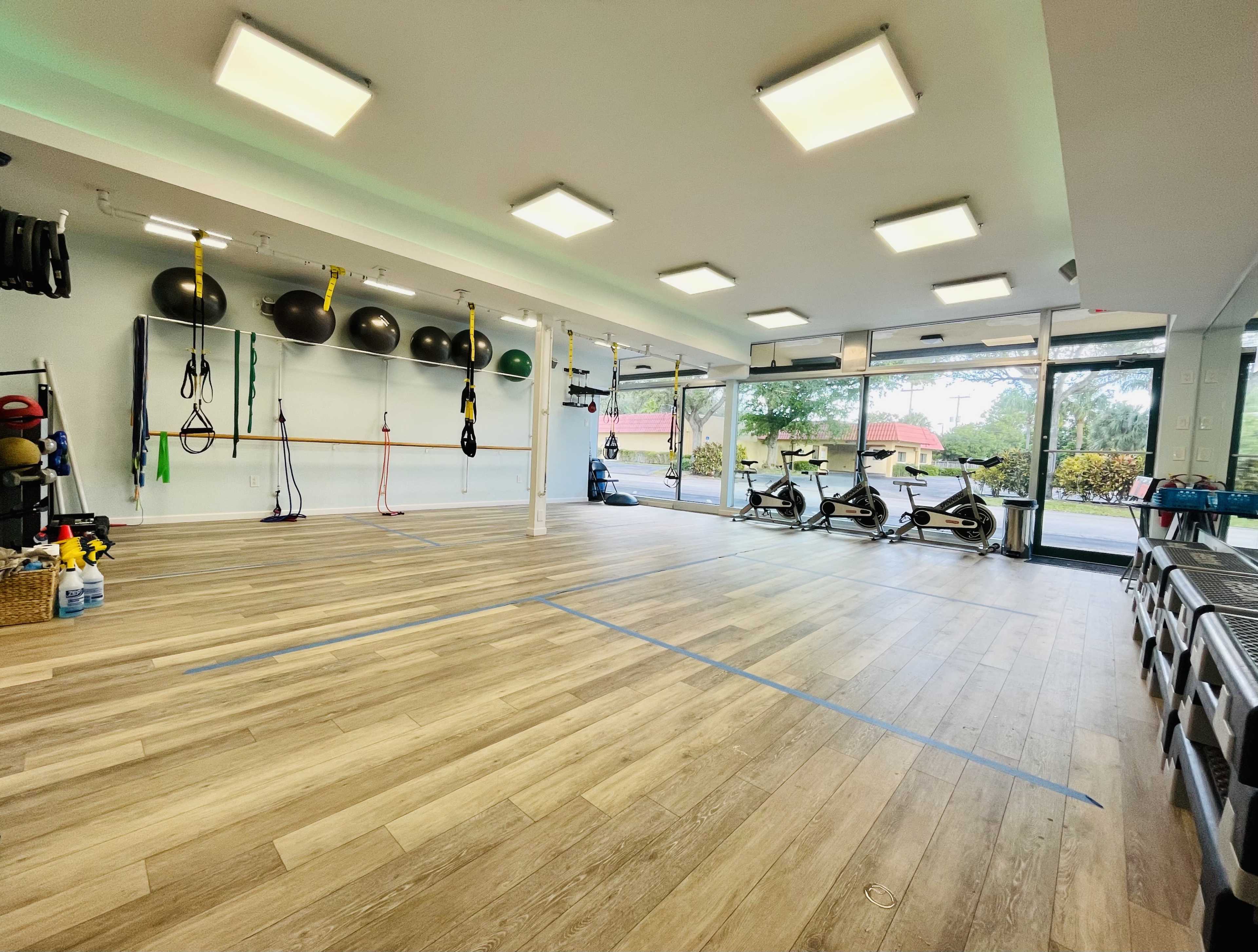 The gym features an open floor area with exercise equipment, fitness balls, resistance bands, and several stationary bikes near large windows.