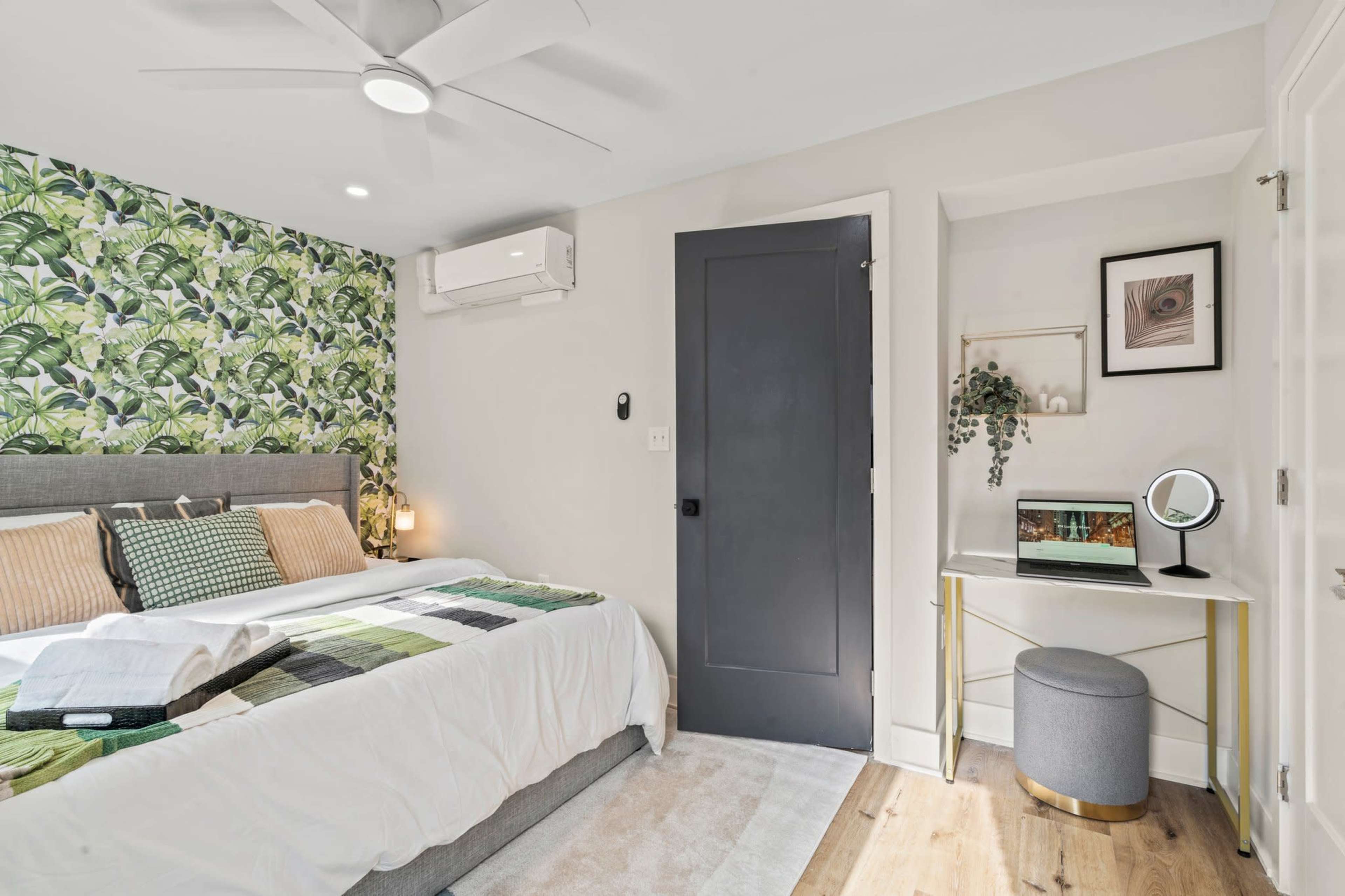 A bedroom with a bed, a small desk and mirror, a closet door, and walls adorned with a leafy wallpaper.
