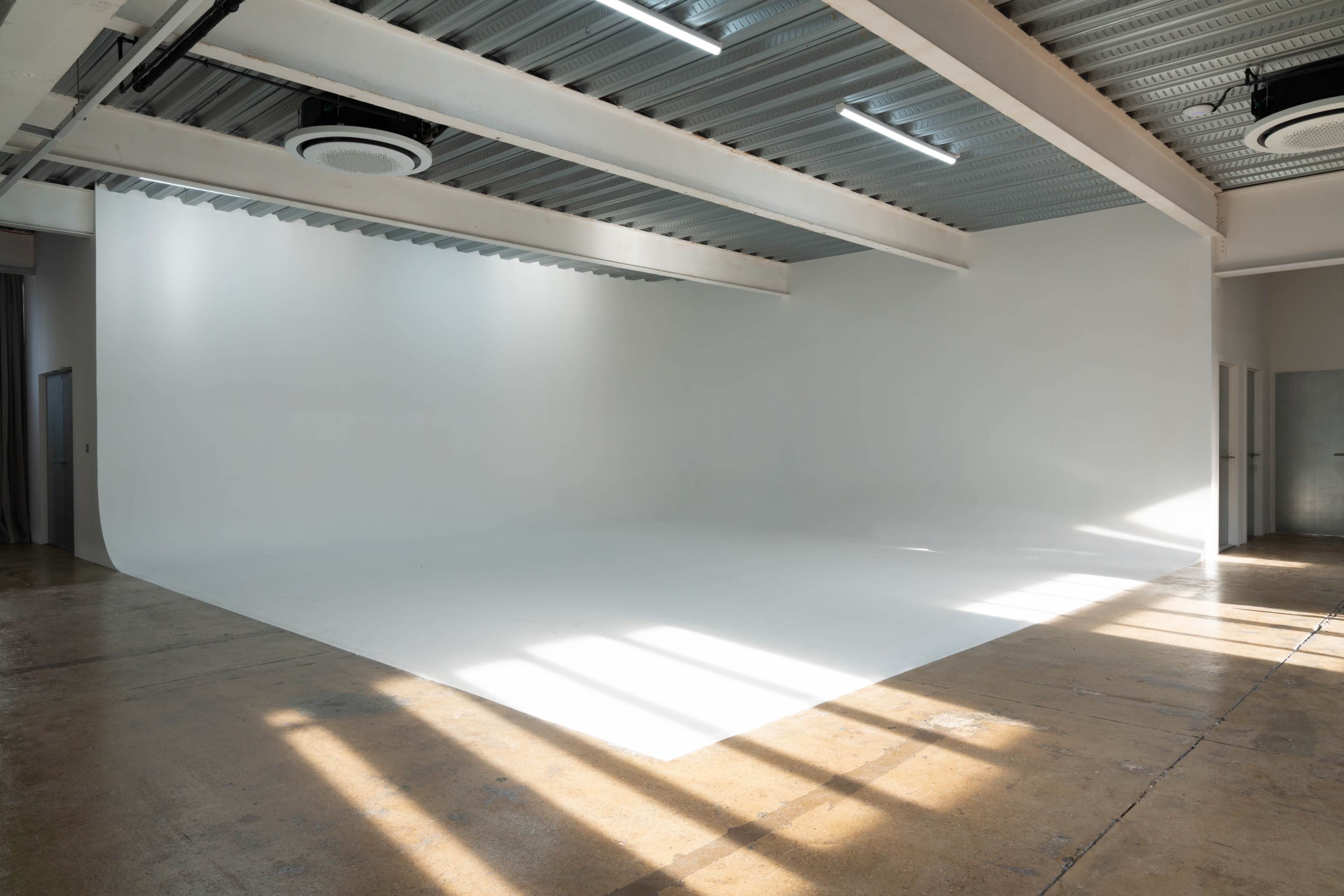 The image shows a spacious, empty room with a white backdrop, concrete flooring, and natural light streaming in through the windows.