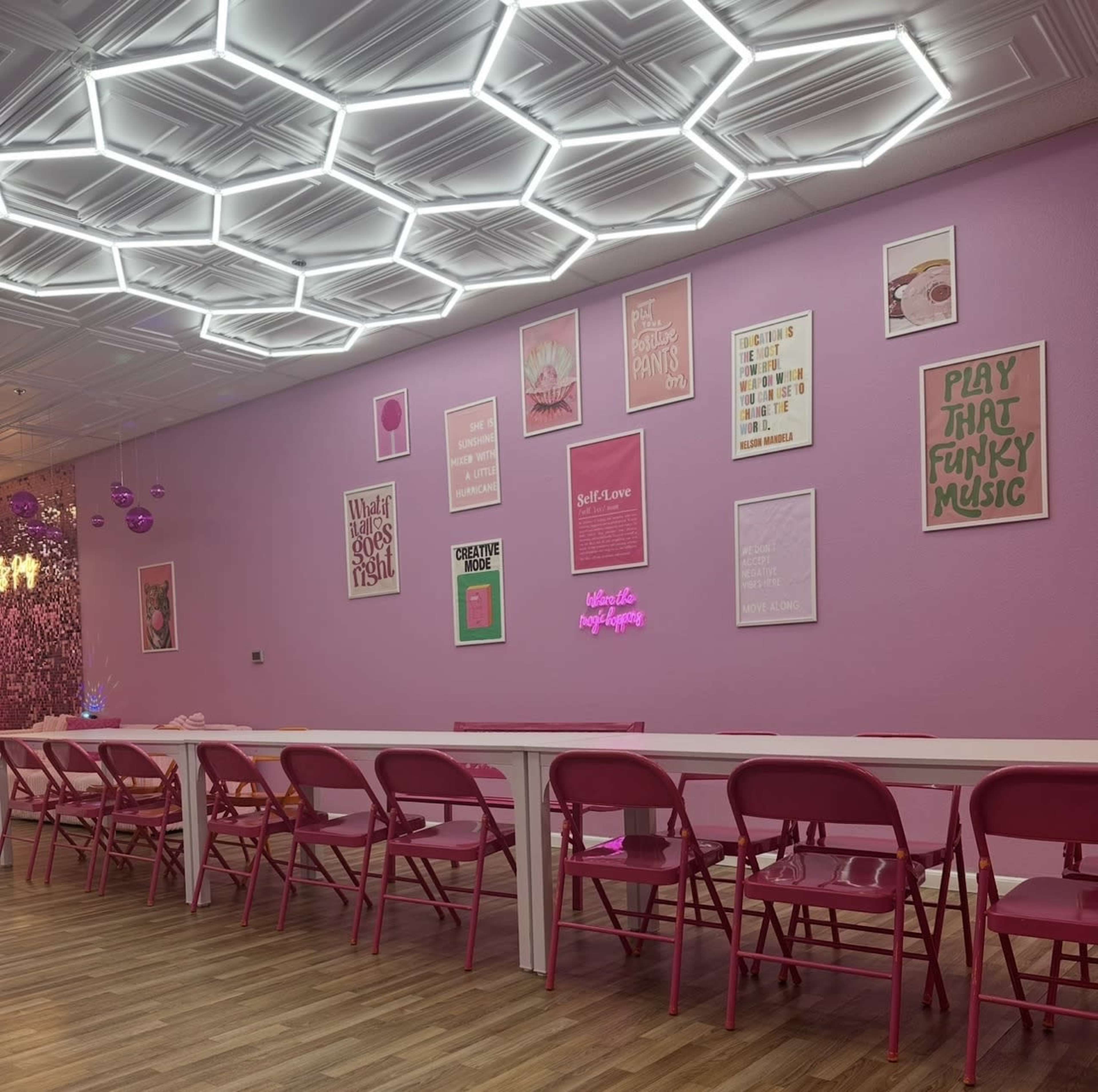 The image shows a brightly colored room with a row of pink chairs and a wall decorated with various framed posters and neon signs.