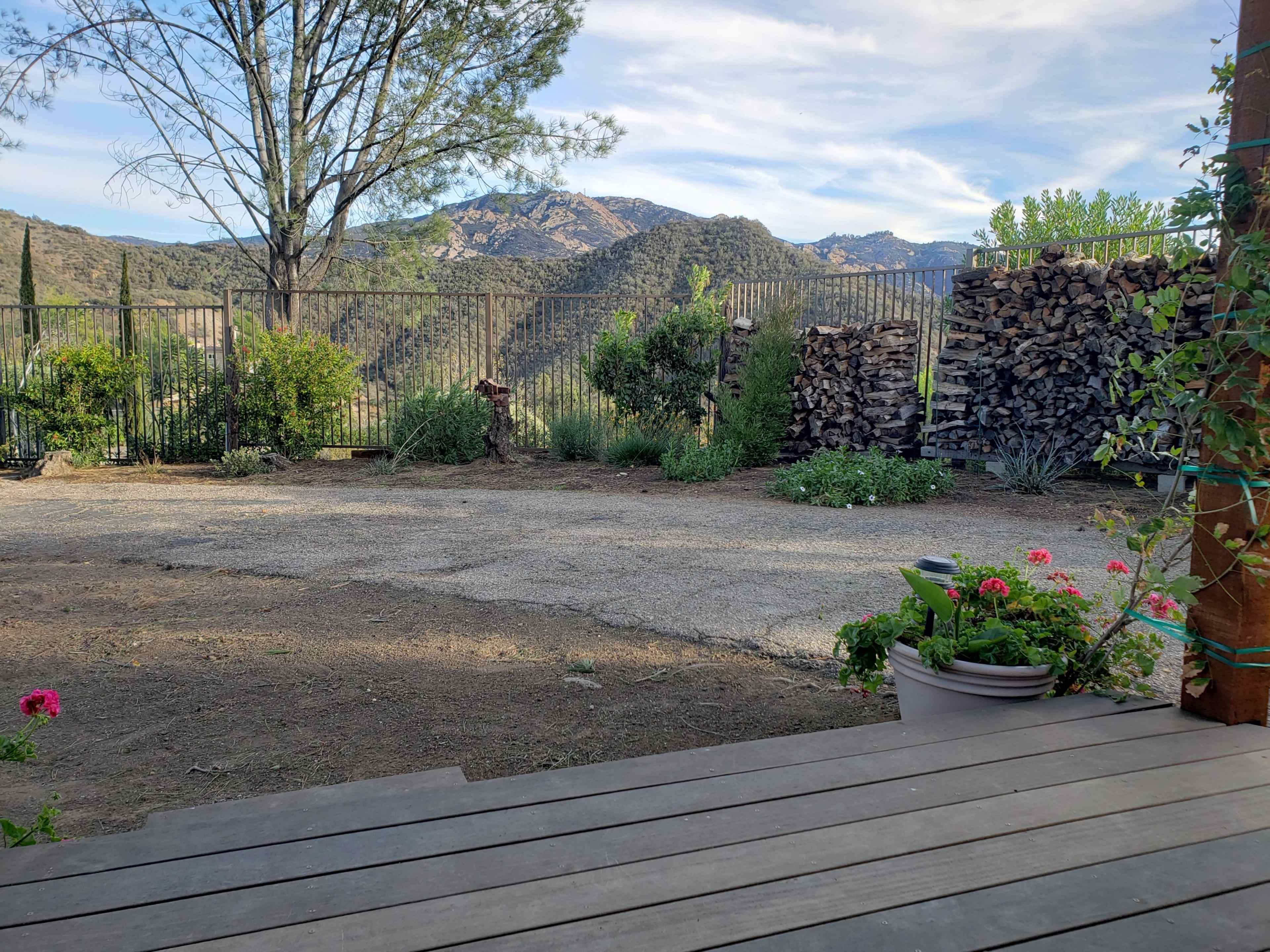 The image shows a gravel area with wooden steps leading down to a yard surrounded by plants, mountains in the background, and a stack of firewood nearby.