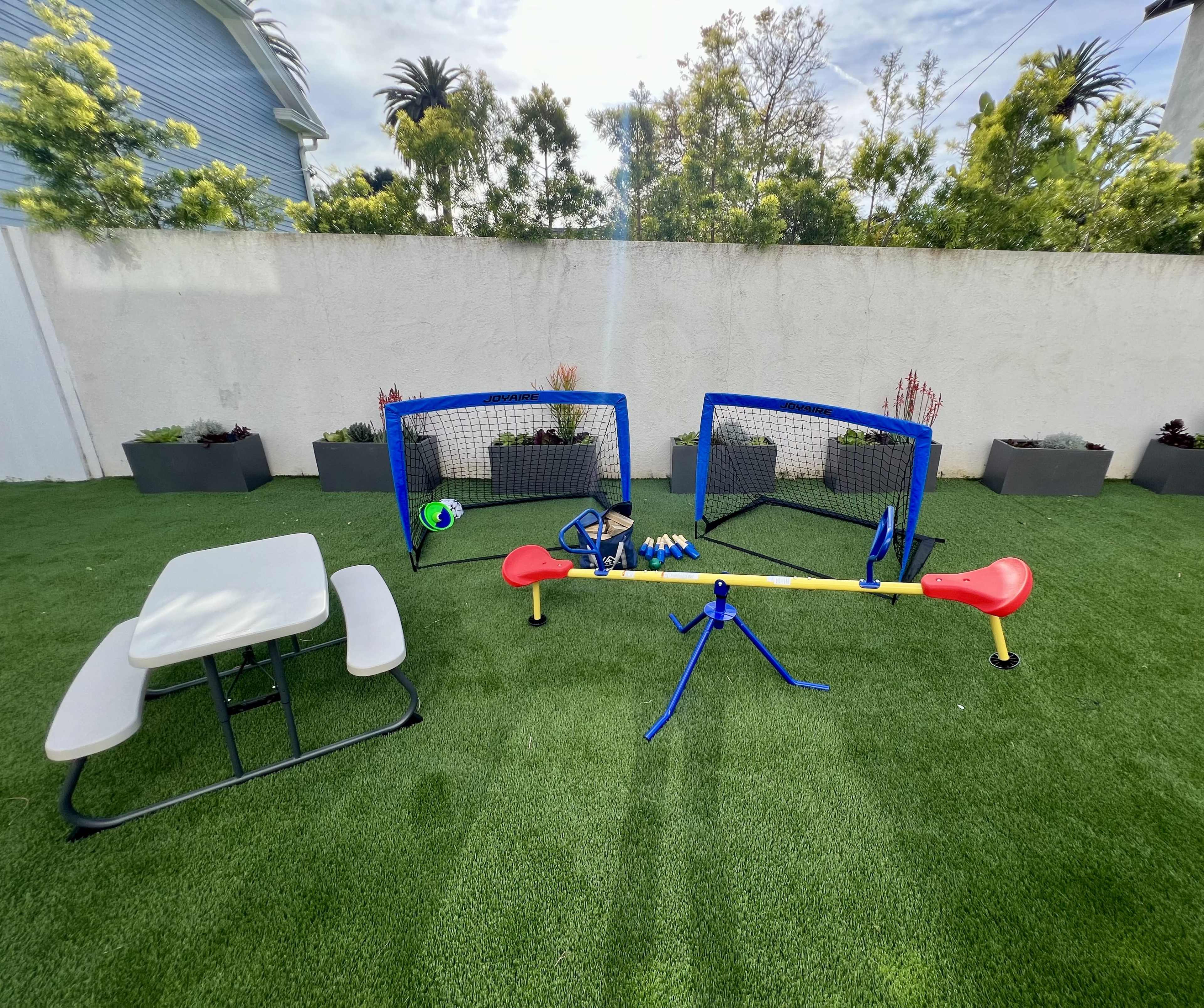 A small outdoor play area with two soccer goals, a ball, and a sports training setup beside a picnic table on green artificial grass.
