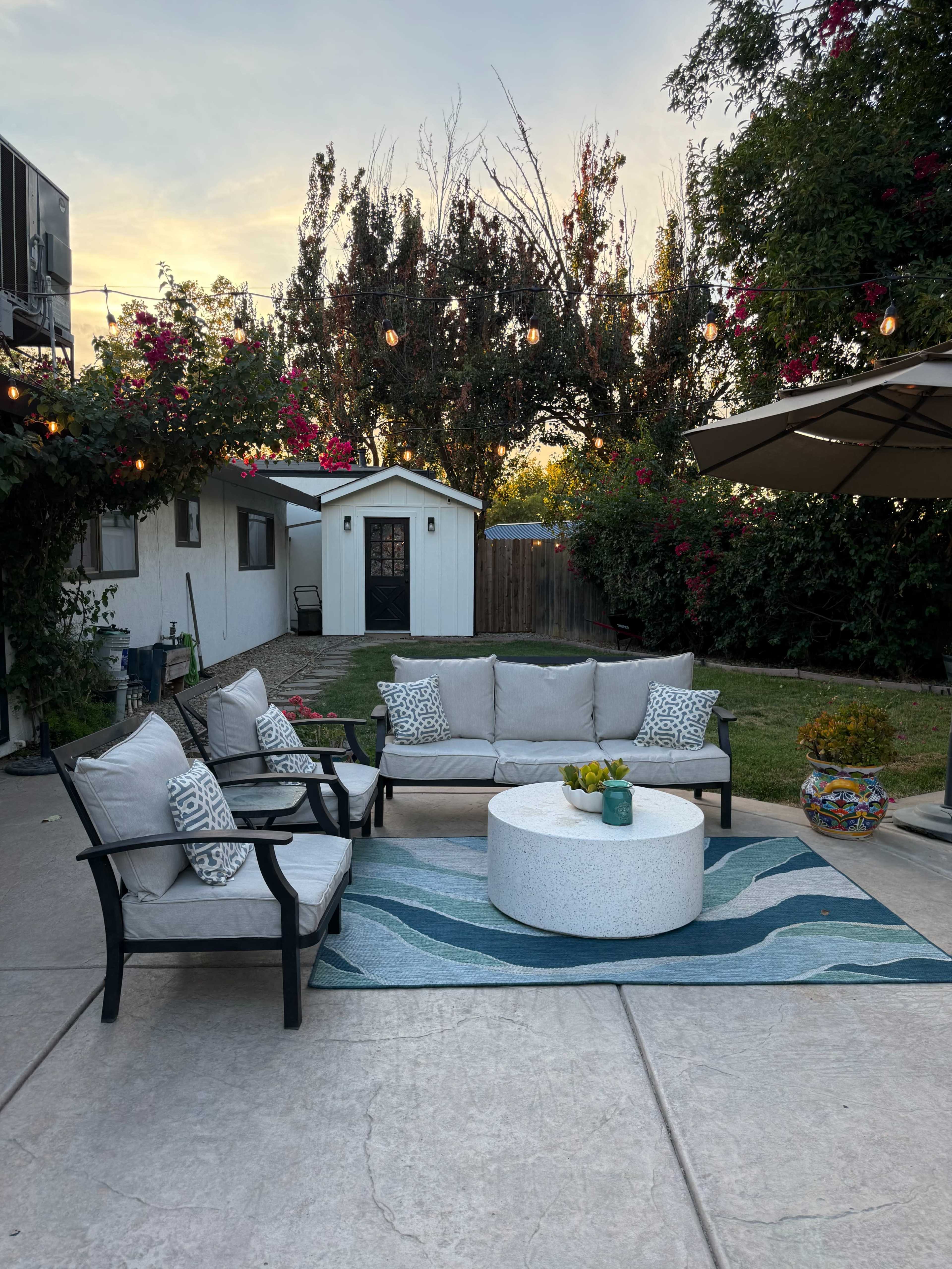 A patio seating area features a circular coffee table surrounded by a sectional sofa and armchairs, with a garden backdrop adorned with flowers and a small shed.