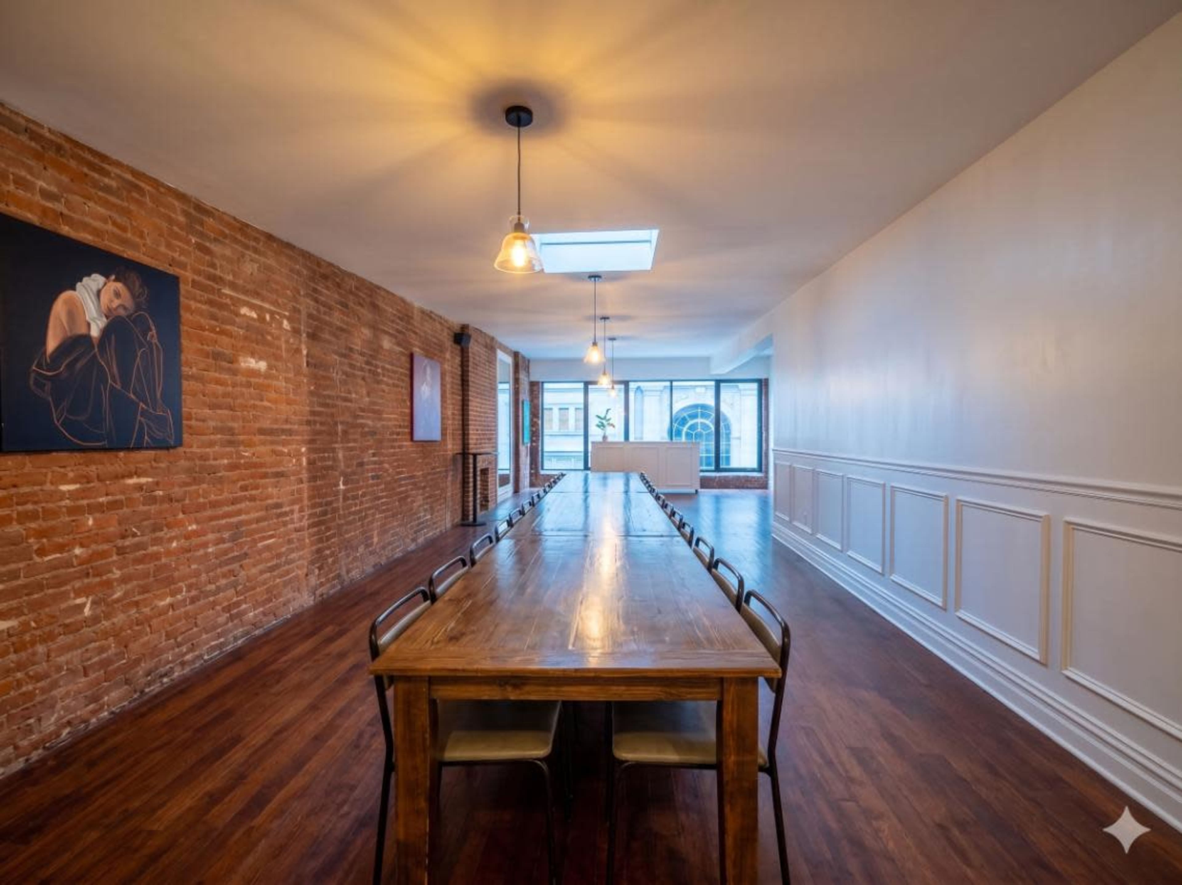 The image shows a spacious room with a long wooden table, exposed brick walls, and large windows allowing natural light.