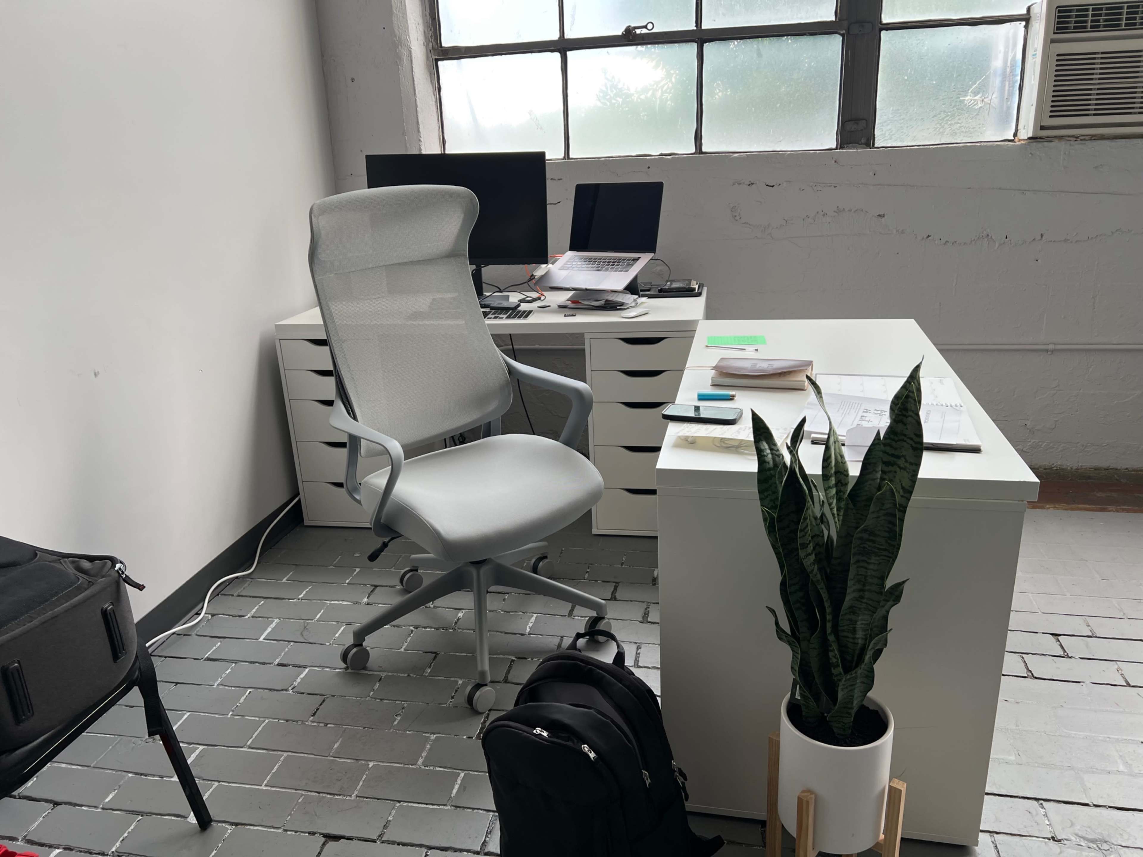 A minimalist office space featuring a white desk with two monitors, a gray office chair, a potted plant, and a backpack on the floor.