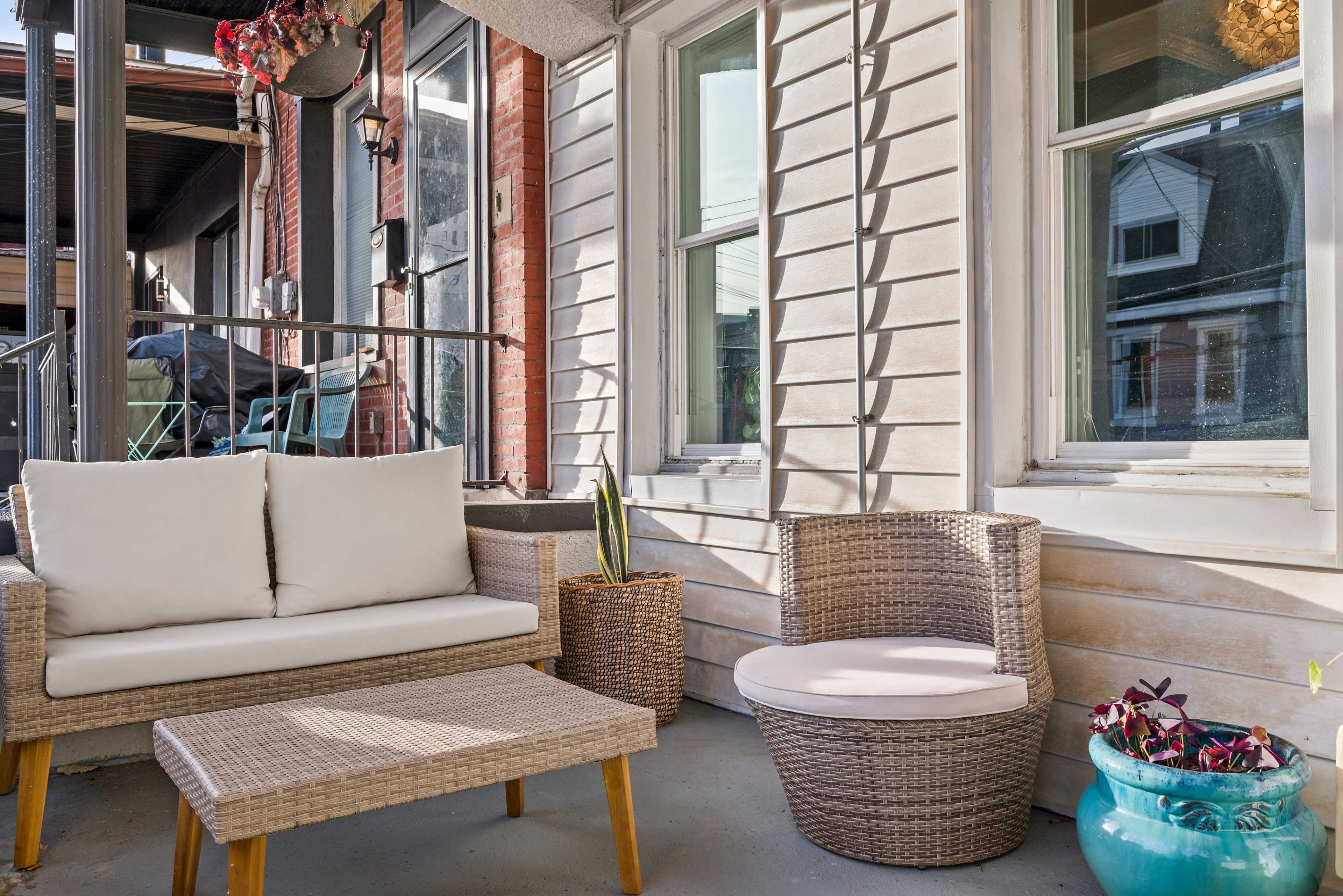A small outdoor seating area featuring a wicker sofa, a side table, and a round chair, surrounded by windows and brick walls.