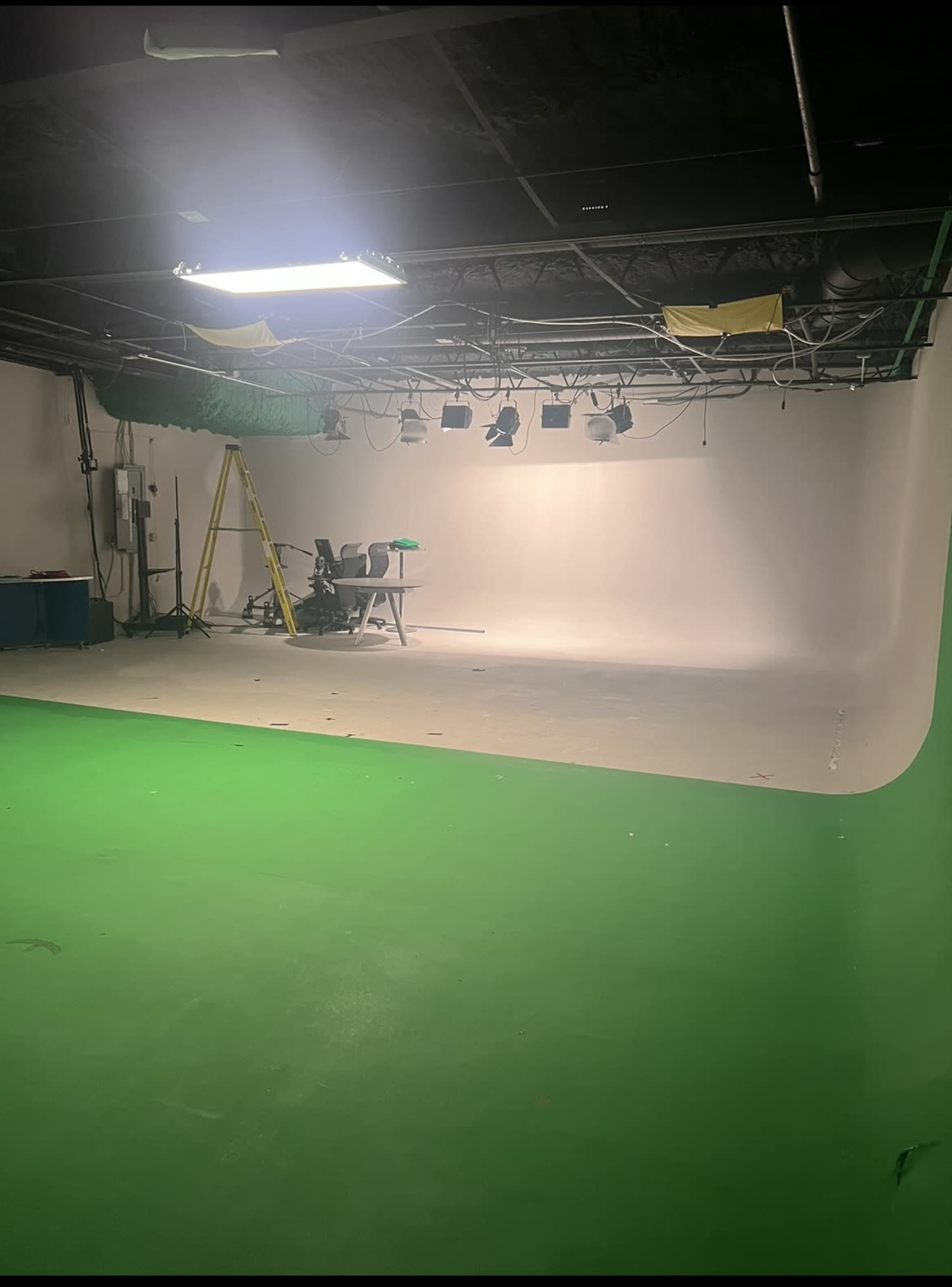 A spacious, empty film studio features a white background, a green floor, a table and chairs, and a ladder against a wall.