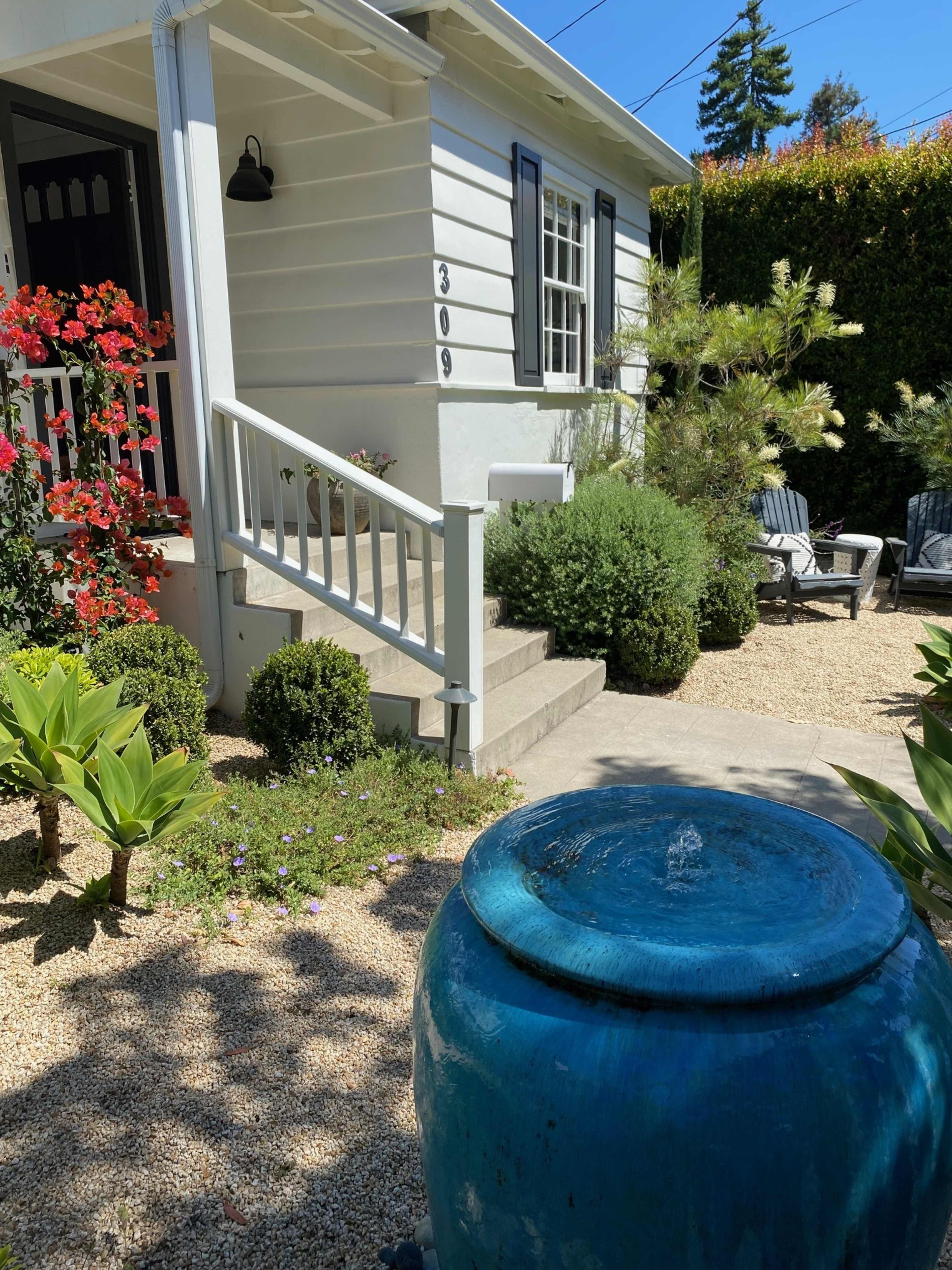 A blue ceramic fountain stands in a landscaped front yard with shrubs and flowering plants beside a white house.