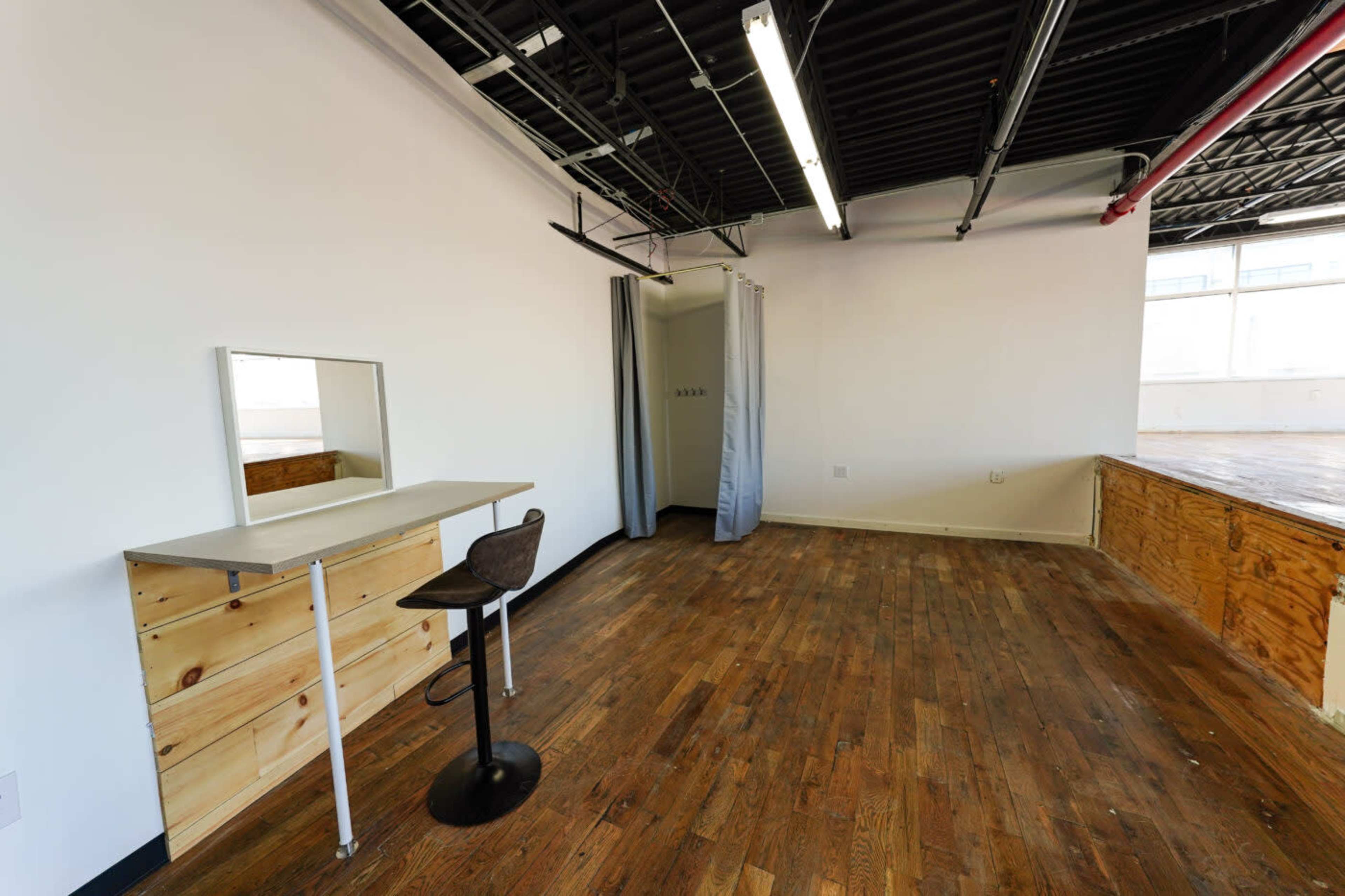 A sparsely furnished room with a wooden floor, a simple desk with a mirror, a stool, and a curtain separating a small area.