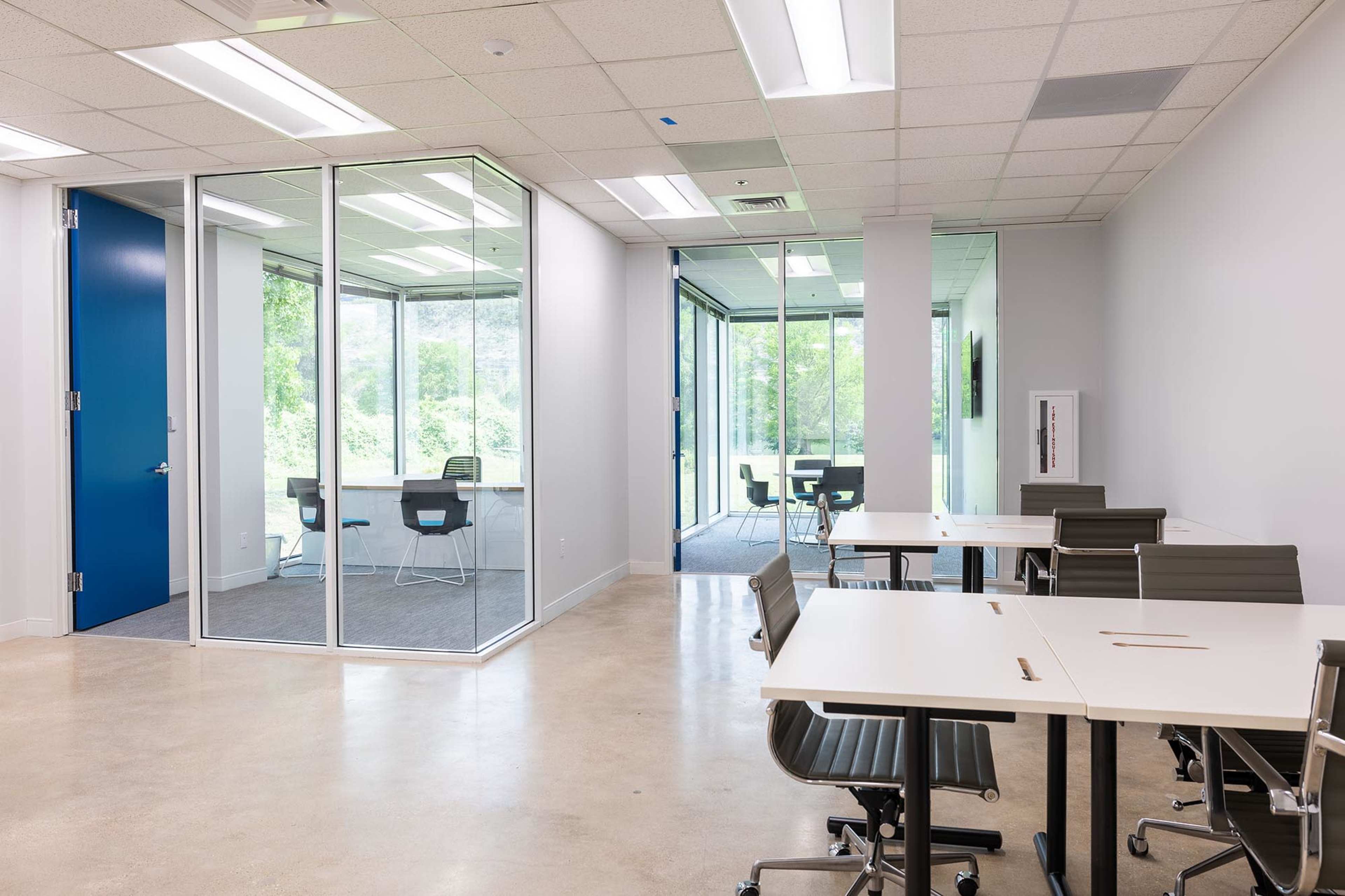 The image shows a modern office space featuring glass-walled meeting rooms, several desks with chairs, and large windows providing a view of greenery.