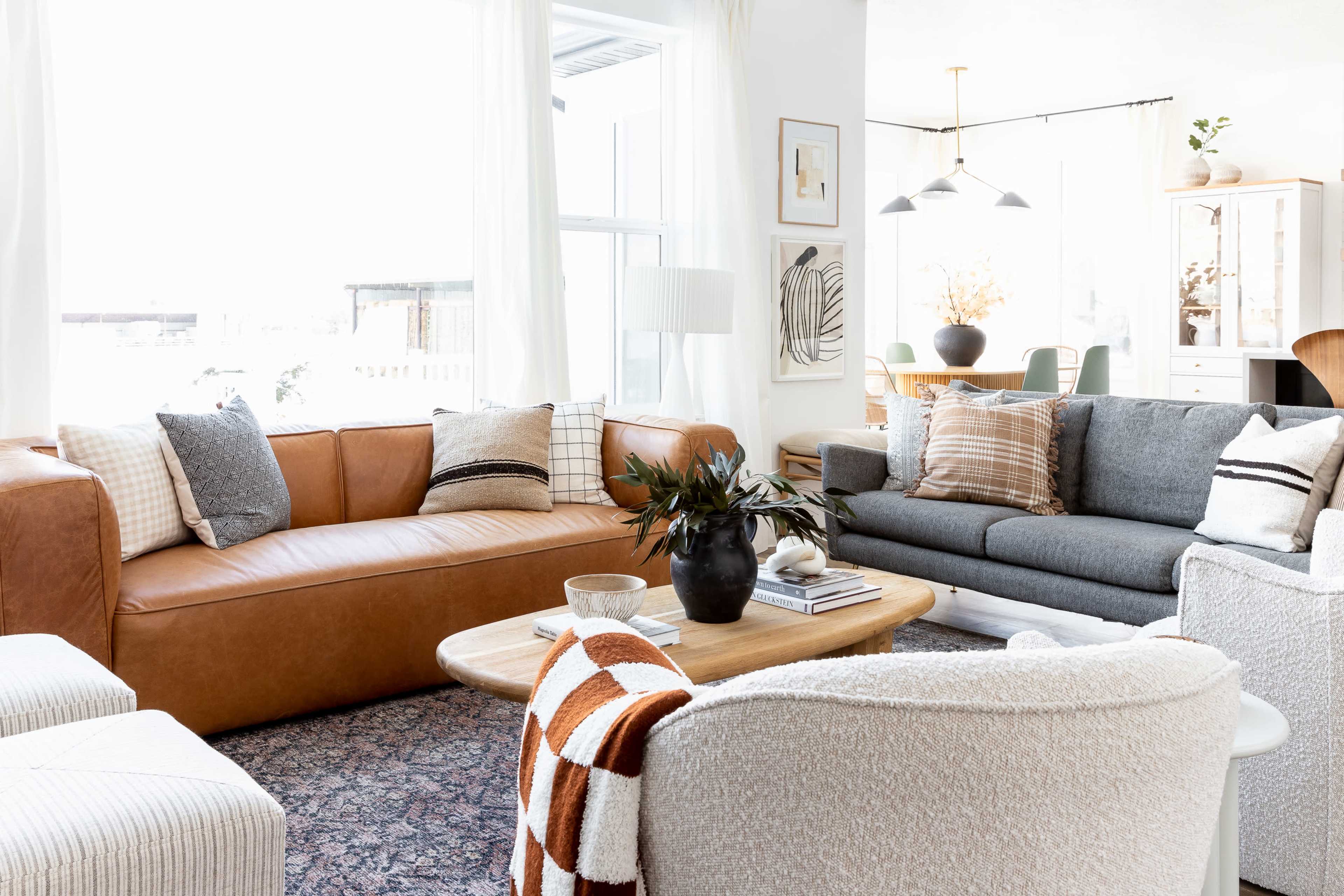 The living room features a brown leather sectional sofa, a gray fabric sofa, and a round wooden coffee table, with large windows allowing natural light to fill the space.