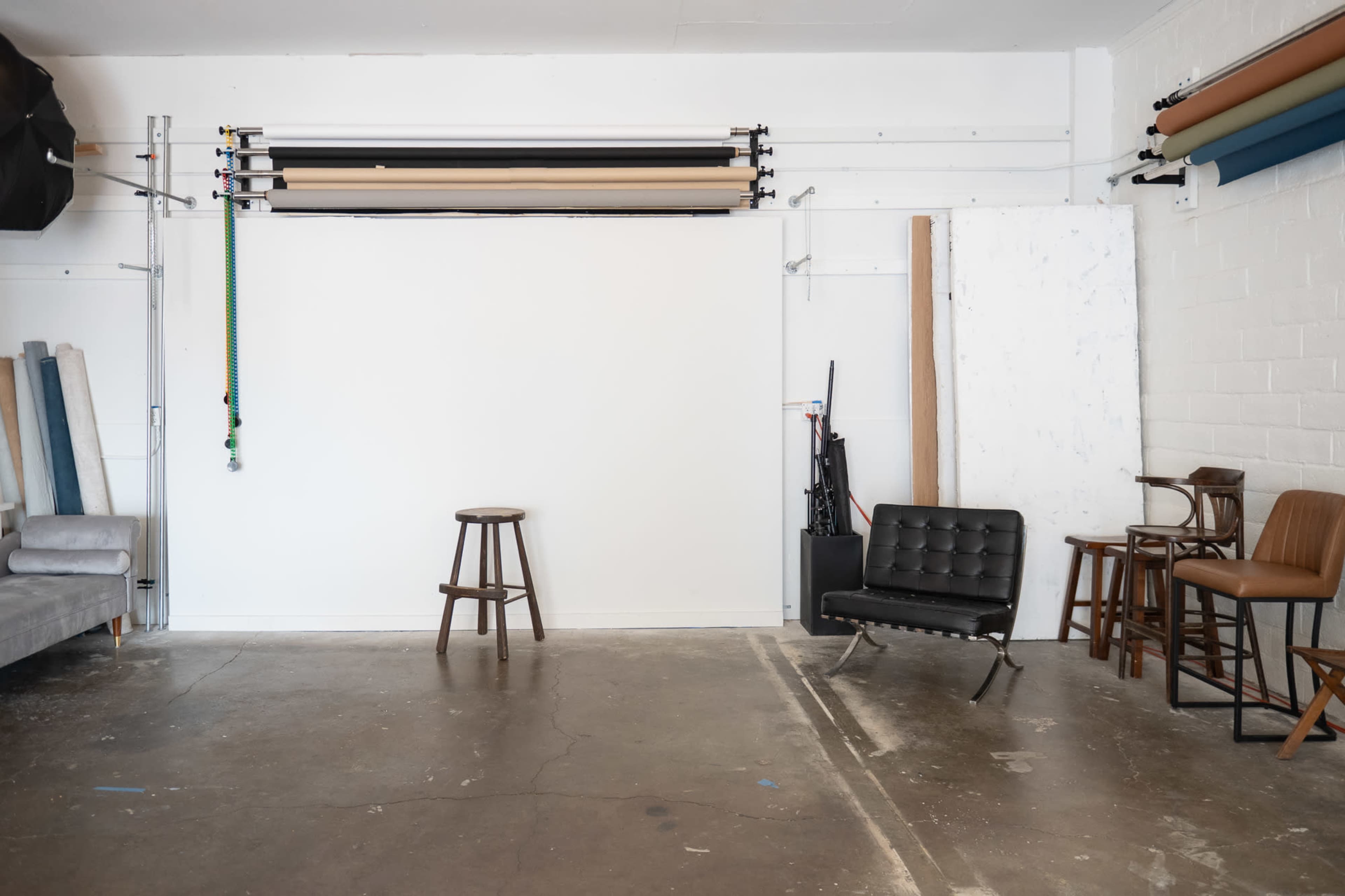 The image shows a photography studio with a blank backdrop, a single stool in the center, and various chairs and backdrops arranged against the walls.