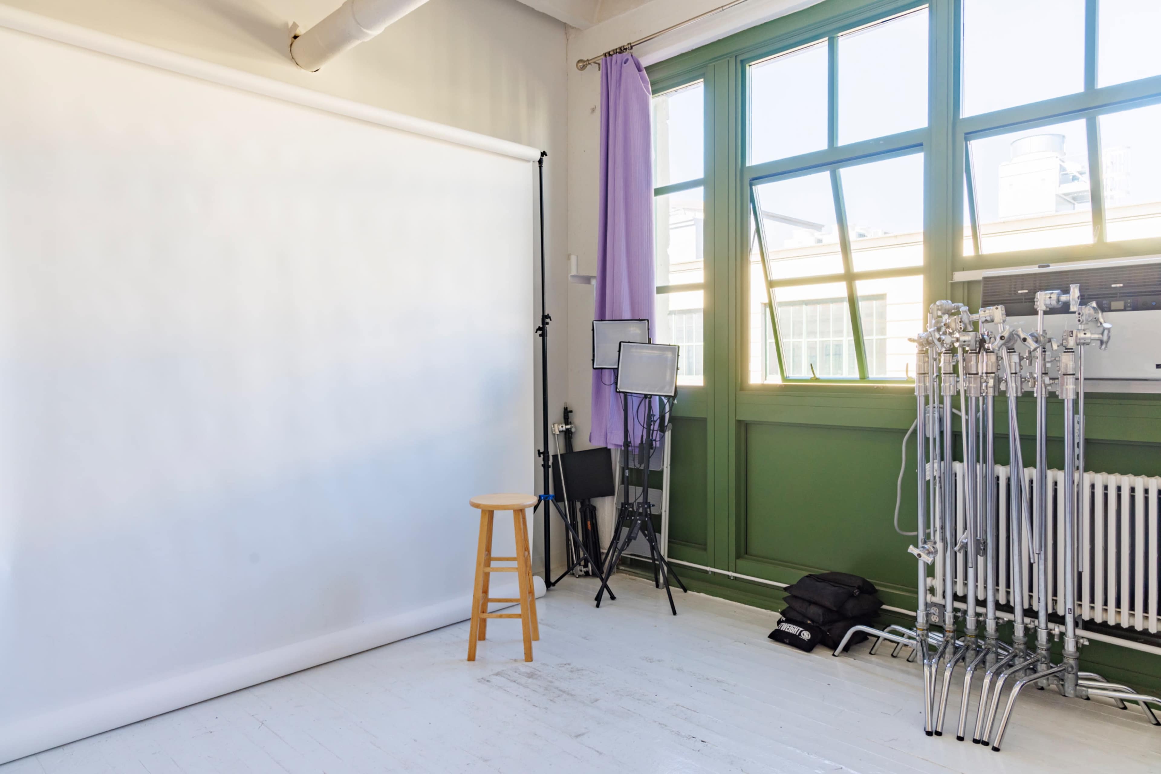 The image shows a photography studio with a white backdrop, a wooden stool, and various lighting equipment.