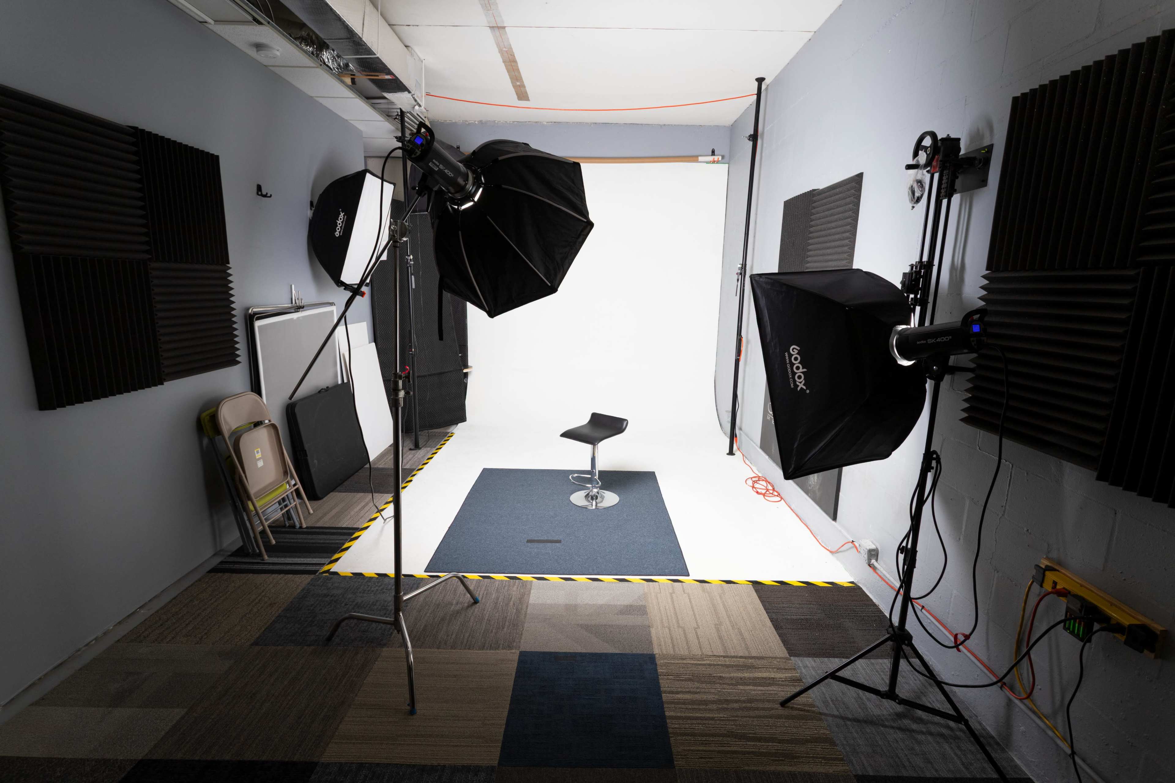 The image shows a photography studio with two light stands and a stool positioned on a light-colored backdrop, surrounded by soundproofing panels and some folding chairs.