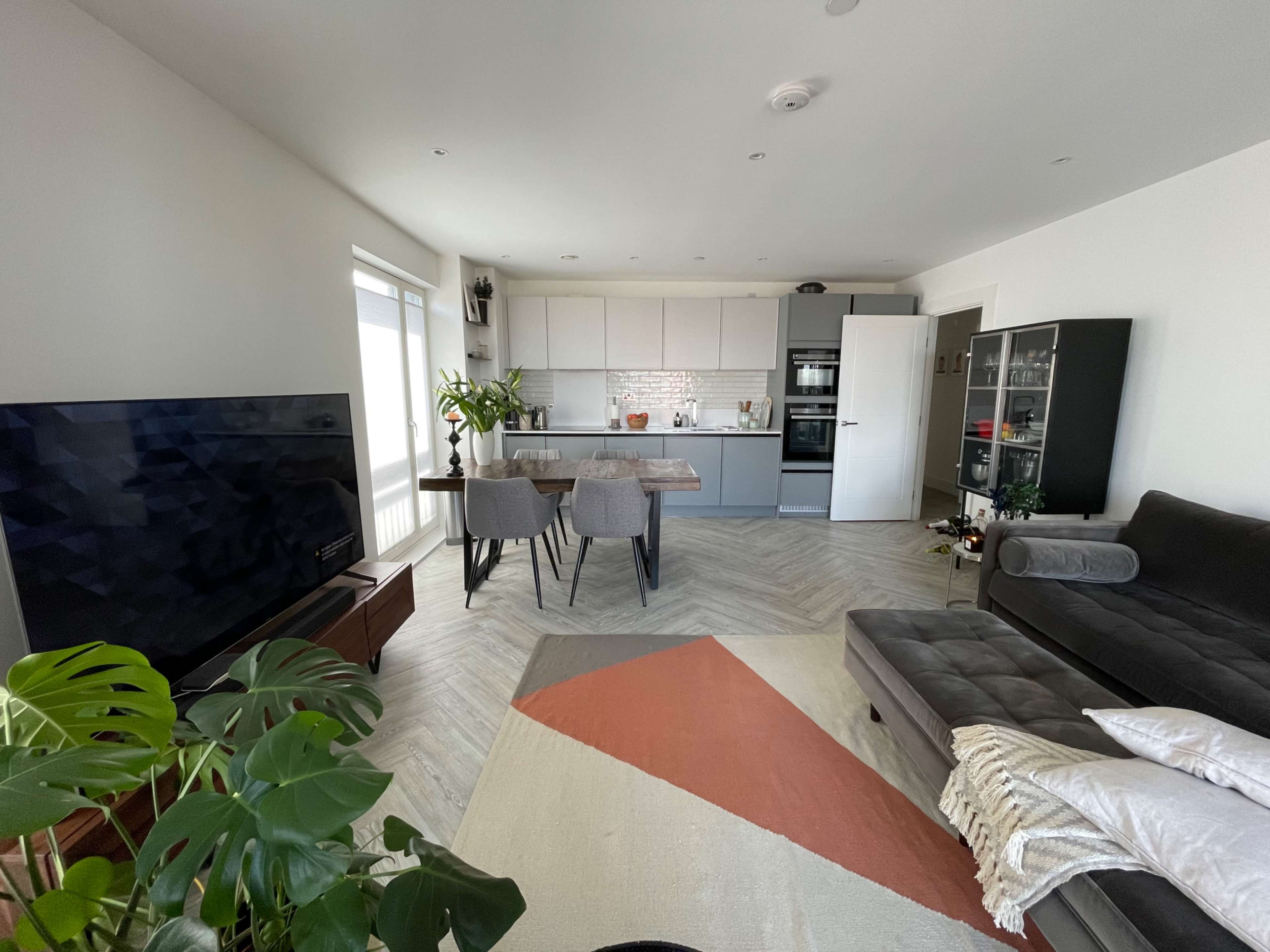 A modern living space features a kitchen area with a dining table, a large TV, a sectional sofa, and potted plants.