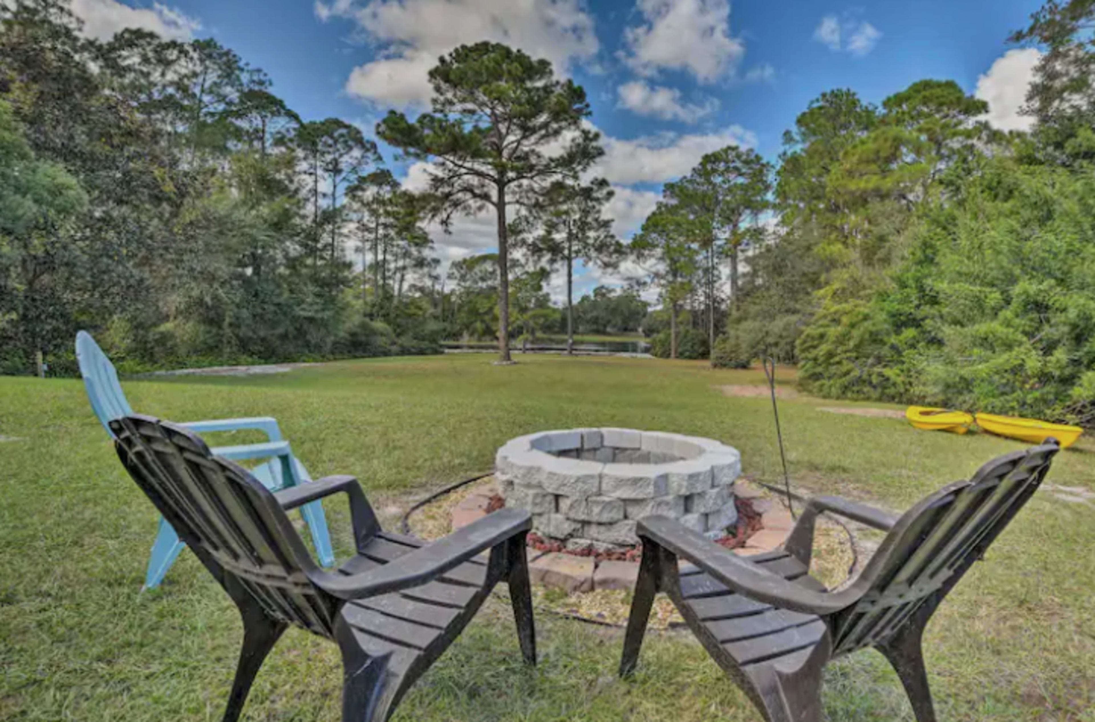 A circular stone fire pit is surrounded by four chairs on a grassy area near a body of water and tall trees.