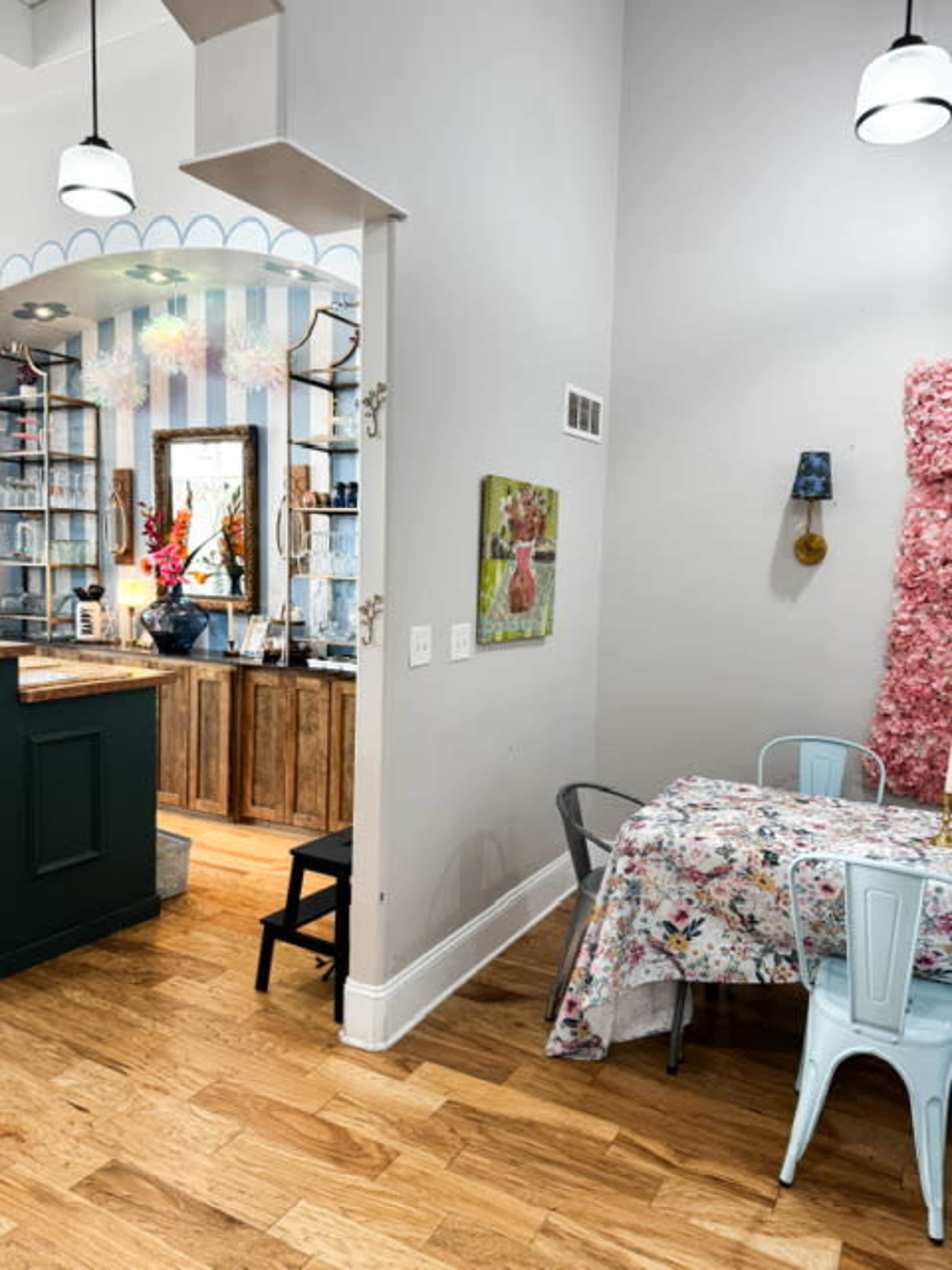 The image shows a cozy dining area with a floral tablecloth and blue metal chairs, leading into a kitchen with wooden cabinets and decorative elements.