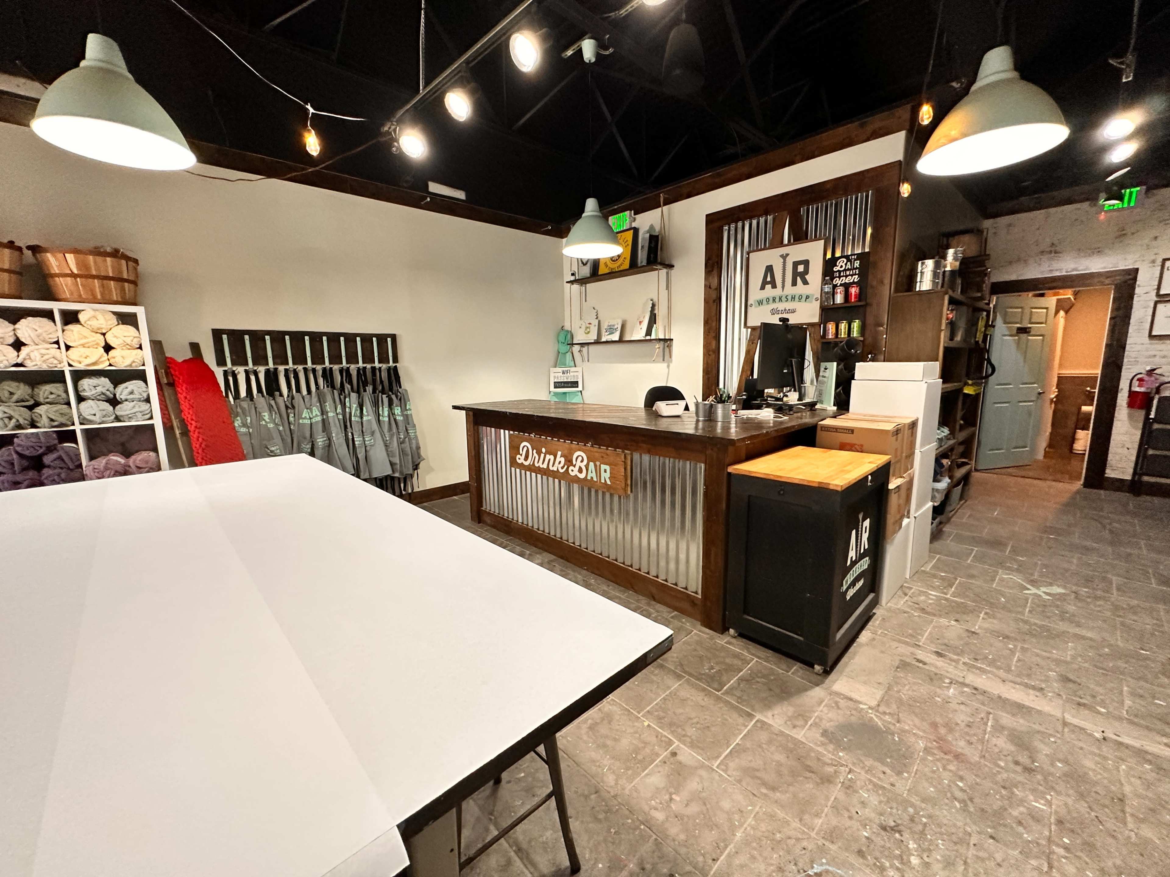 The image shows a well-lit bar area with a wooden counter, shelves filled with items, and a wall displaying folded towels in a neatly organized space.