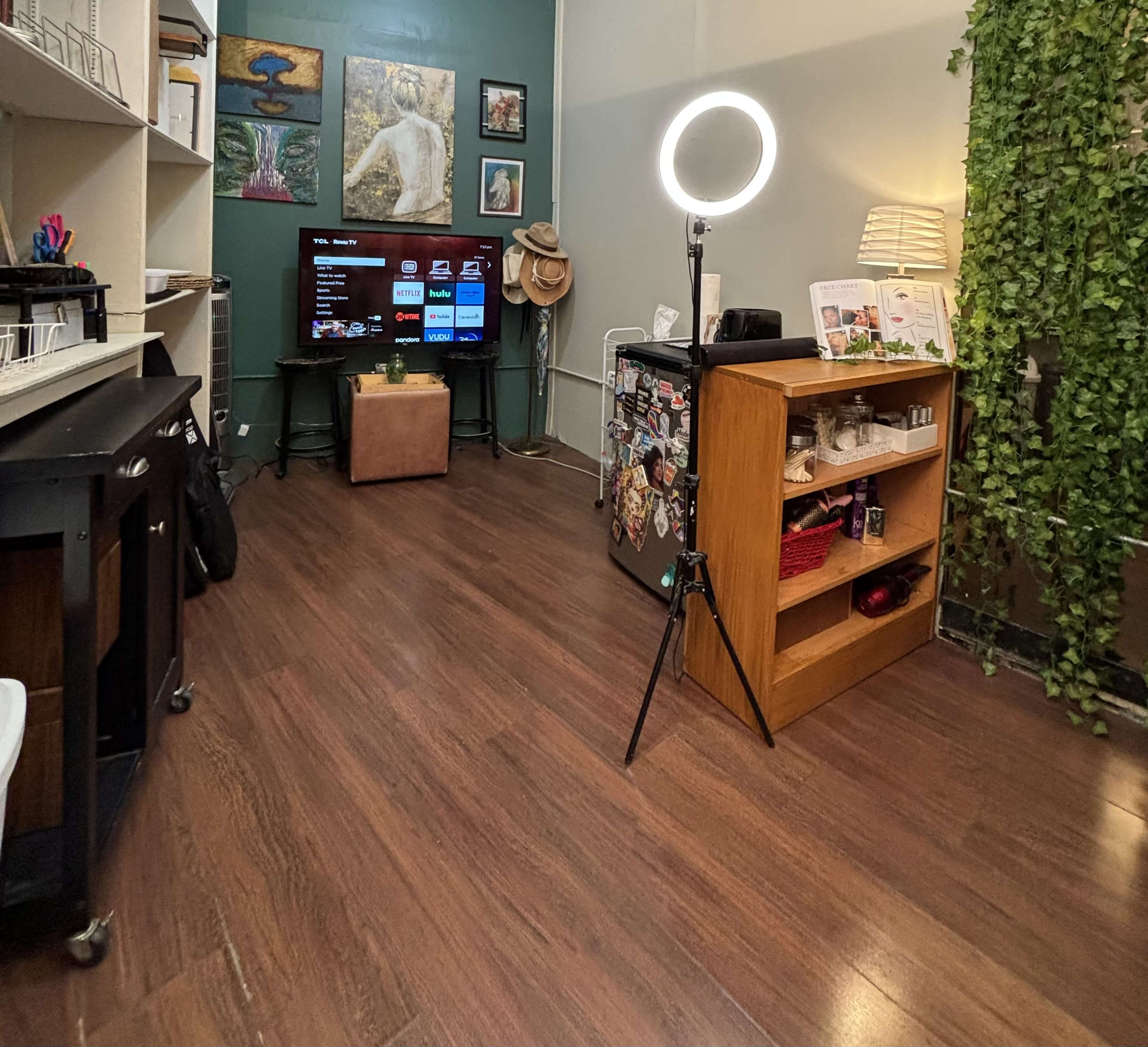 A room with wooden flooring, a television on a stand, a shelf with items, a small seating area, and a ring light on a tripod.