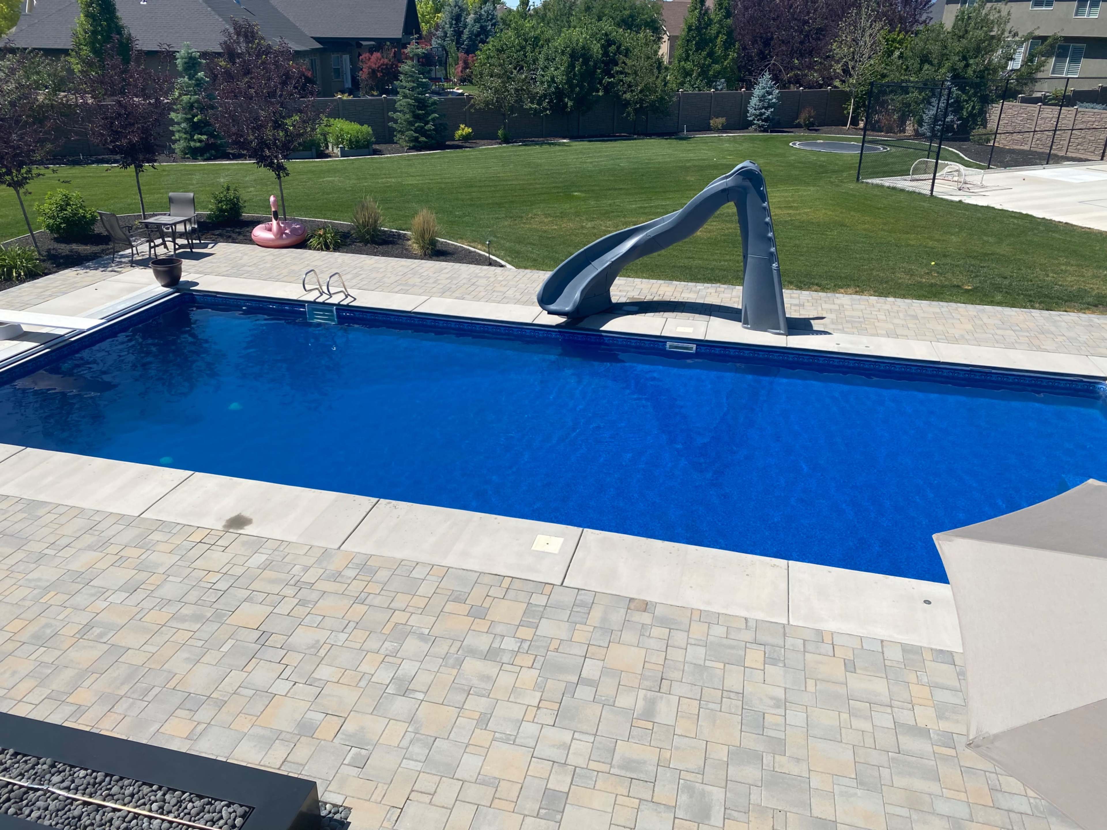 A rectangular swimming pool with a gray slide, surrounded by landscaped lawn and patio areas featuring seating and a fire pit.