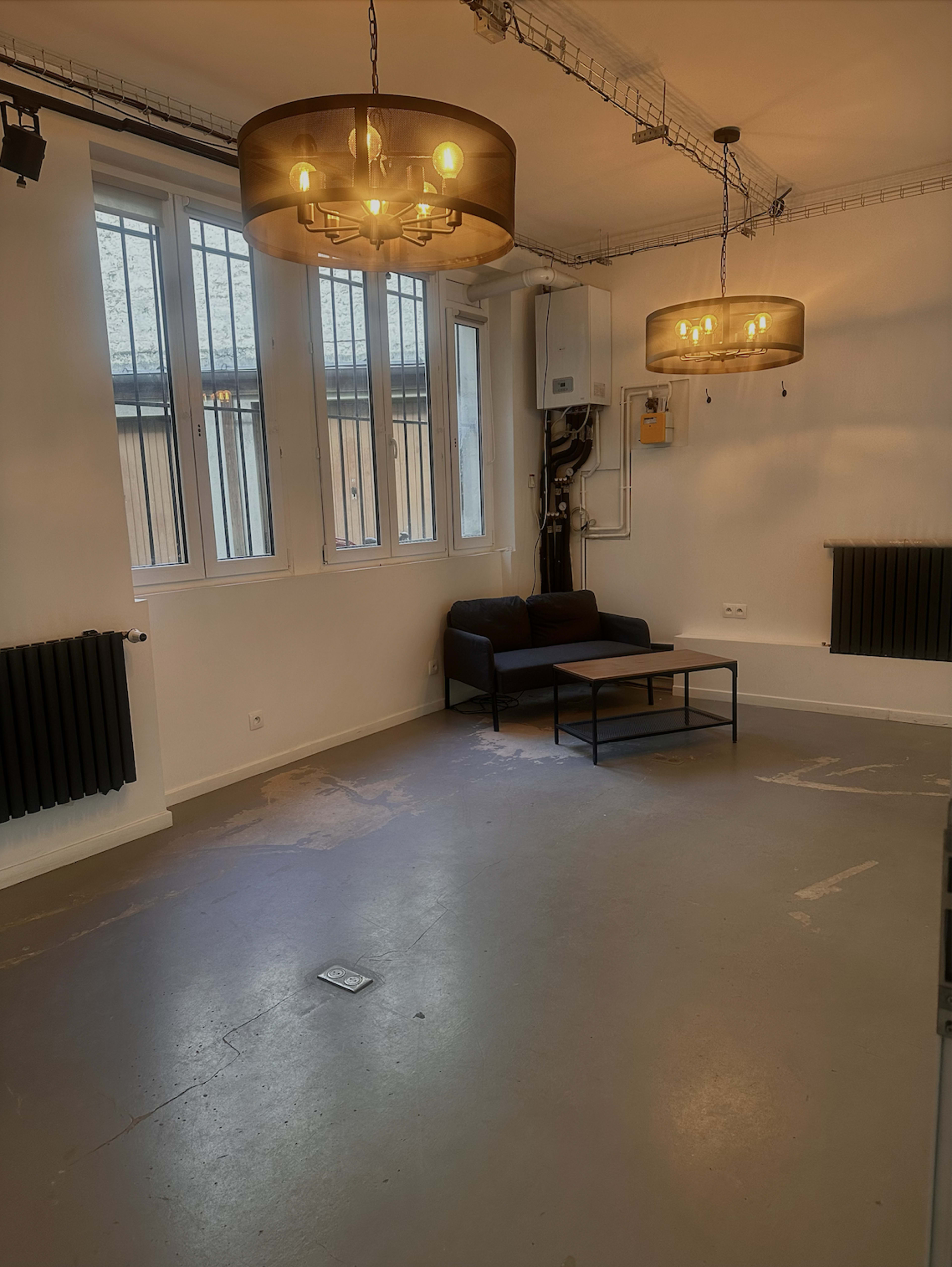 The image shows a spacious, well-lit room featuring a black couch and a coffee table under two hanging light fixtures, with large windows allowing natural light.