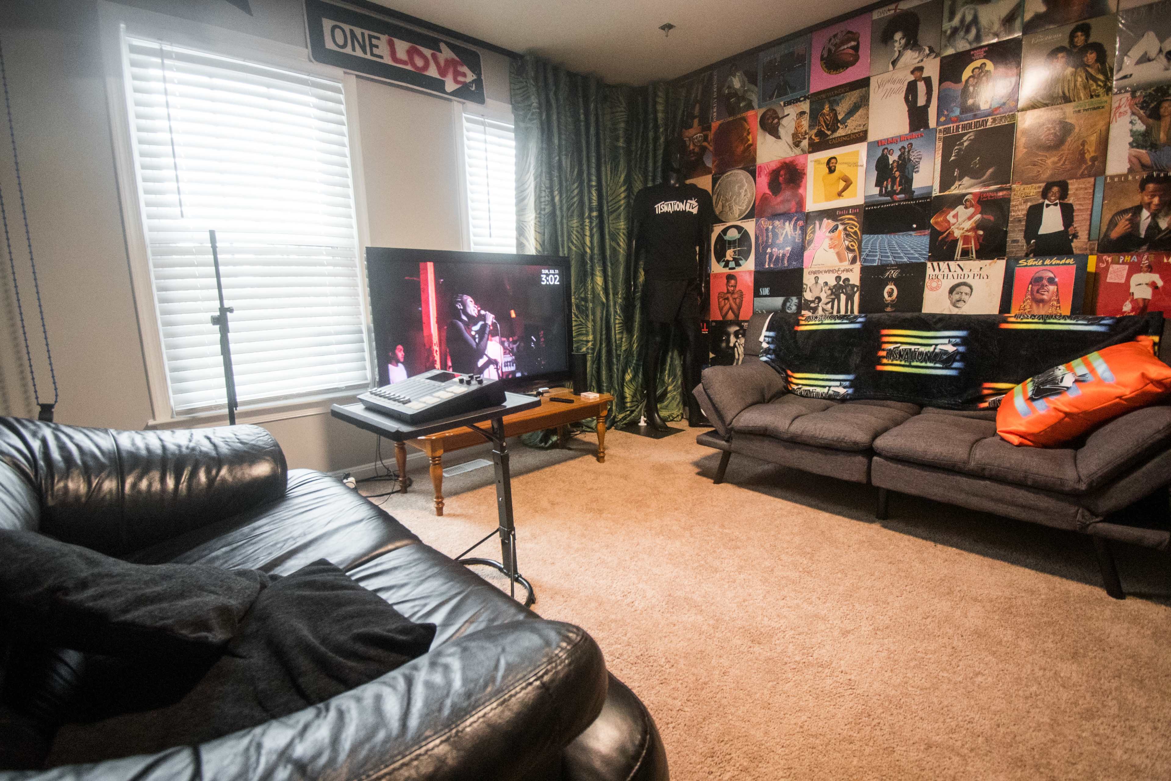 The room features a black leather couch, a television displaying a performer, and a wall covered with colorful album covers and posters.