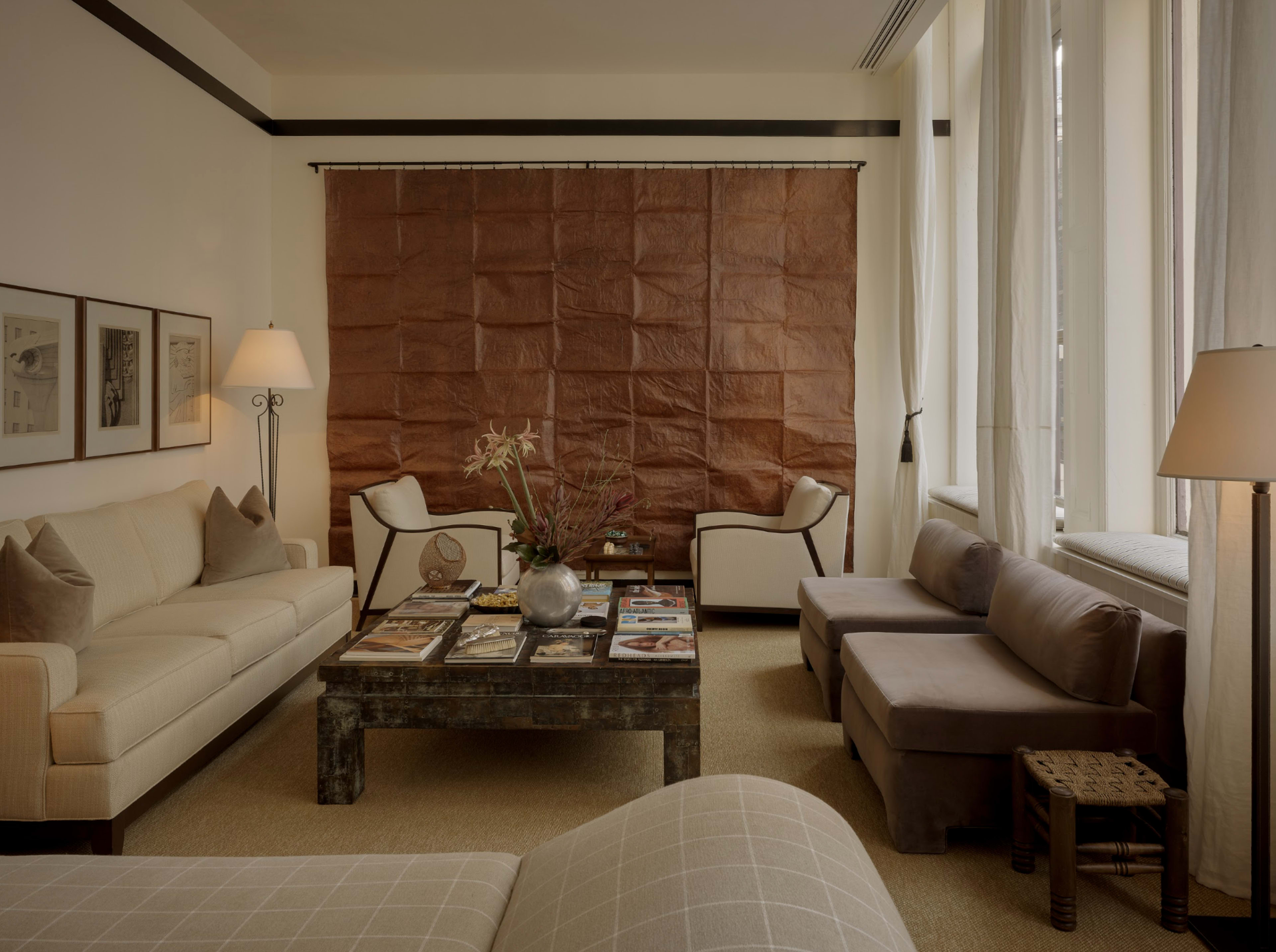 The image depicts a stylish living room featuring a beige sofa, two armchairs, a coffee table with magazines, and a textured brown wall hanging.