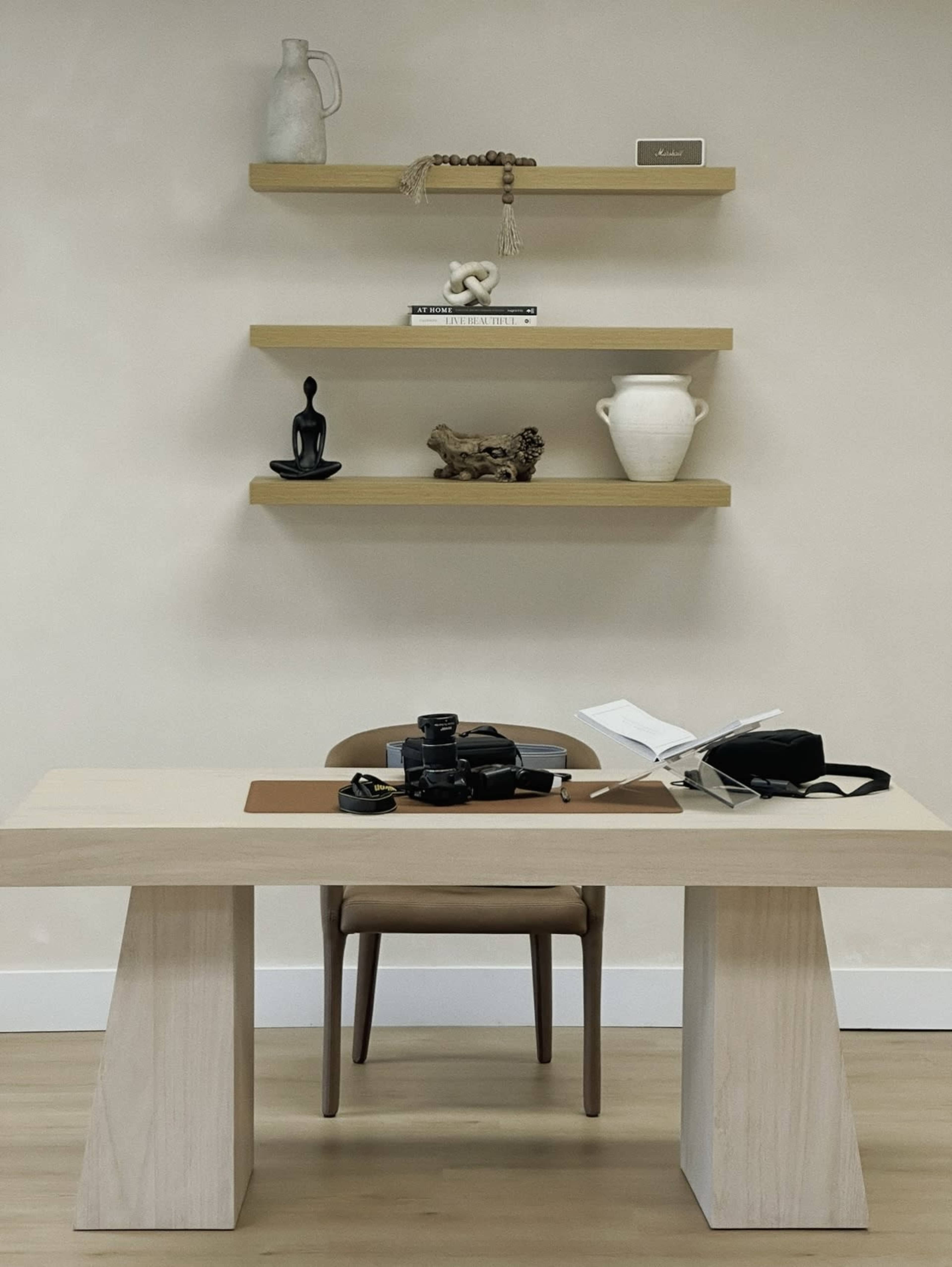 A minimalist workspace features a wooden table, a chair, and shelves displaying various decorative items and a camera.