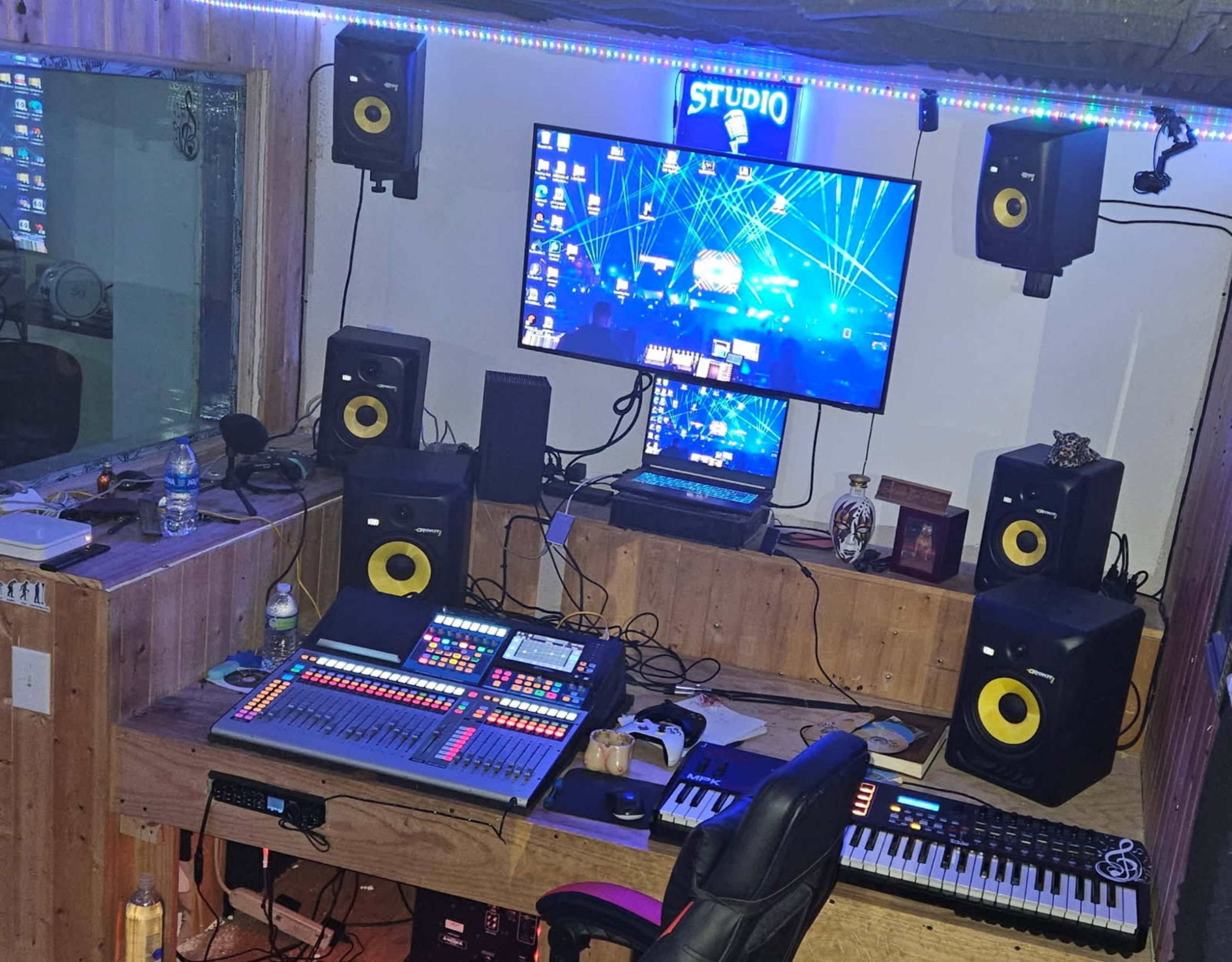 A music studio is set up with a computer, multiple speakers, a mixing console, a keyboard, and colorful LED lighting.