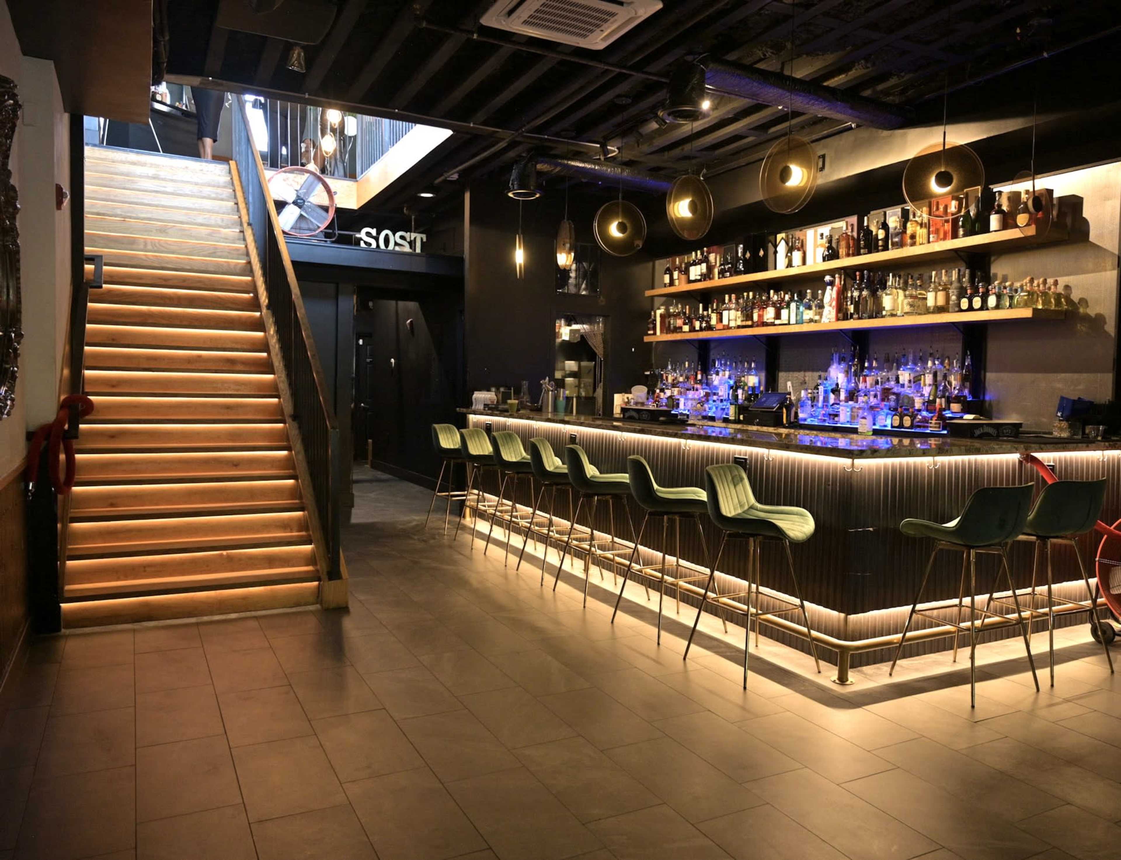 A modern bar features a sleek counter with illuminated bottles and high stools, adjacent to a wooden staircase leading to an upper level.