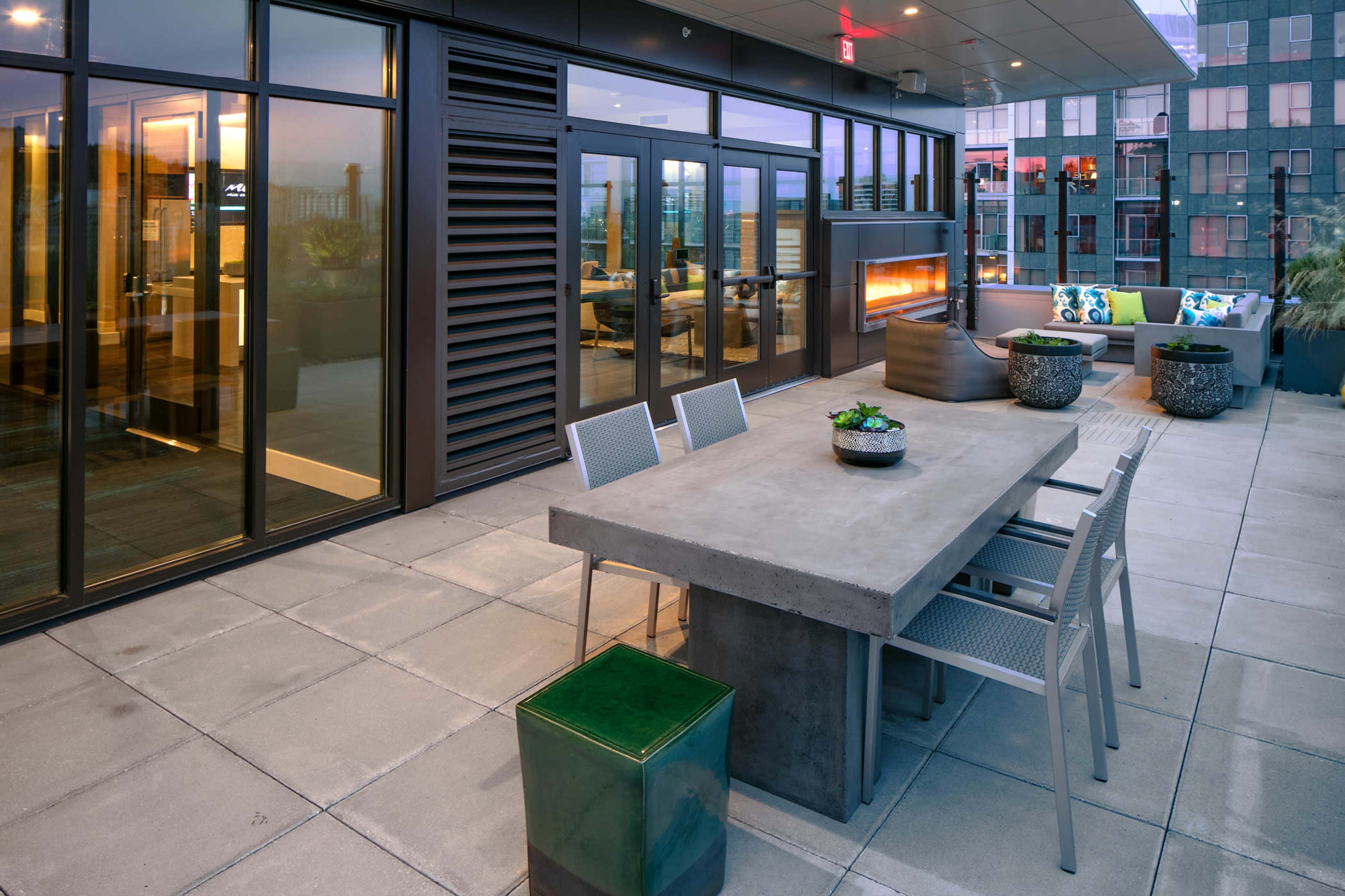 A modern rooftop terrace with a concrete dining table, metal chairs, and an outdoor fireplace, surrounded by glass sliding doors and potted plants.