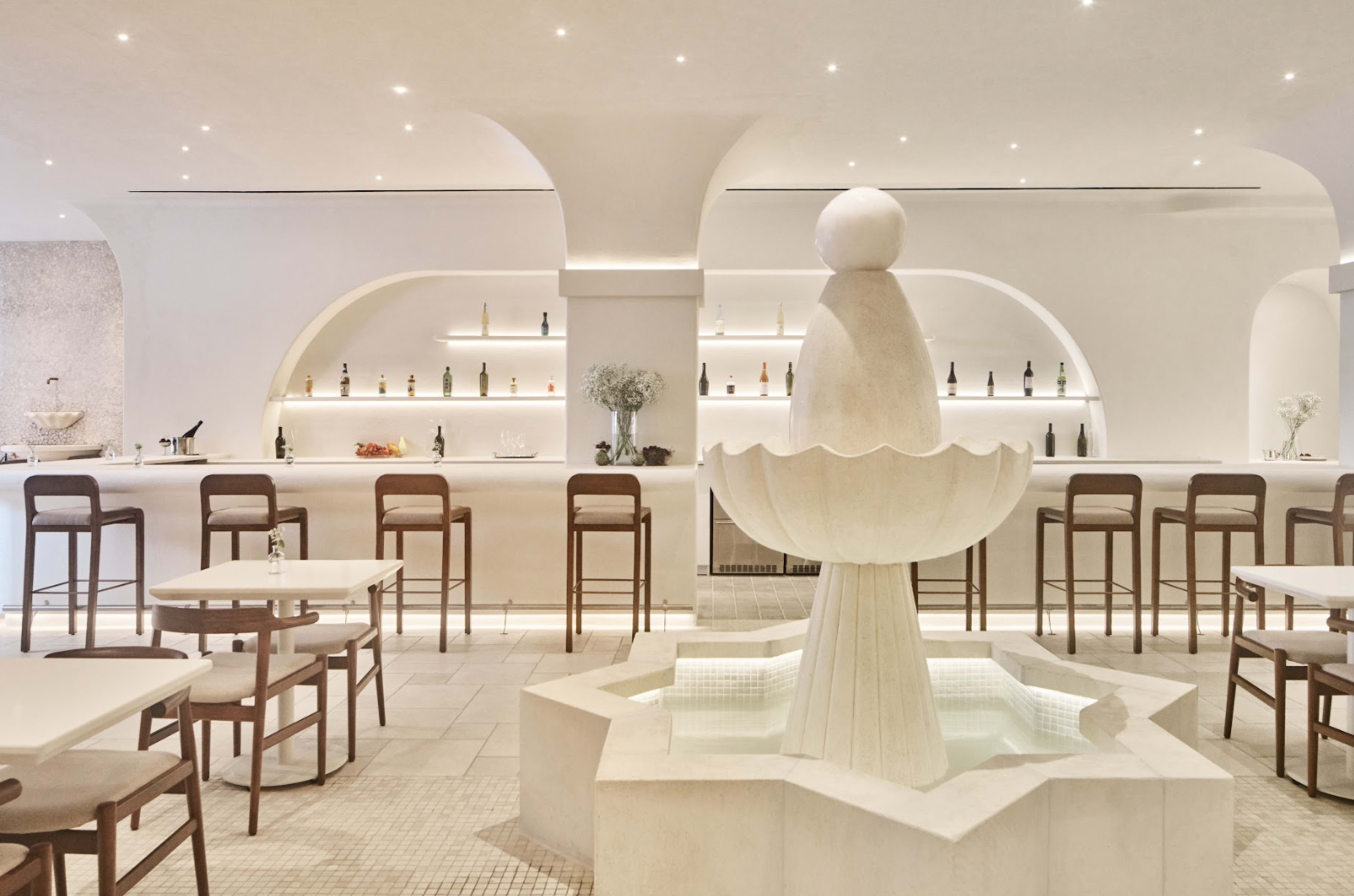 A modern restaurant features a central stone fountain surrounded by white bar stools and tables against a minimalist backdrop.