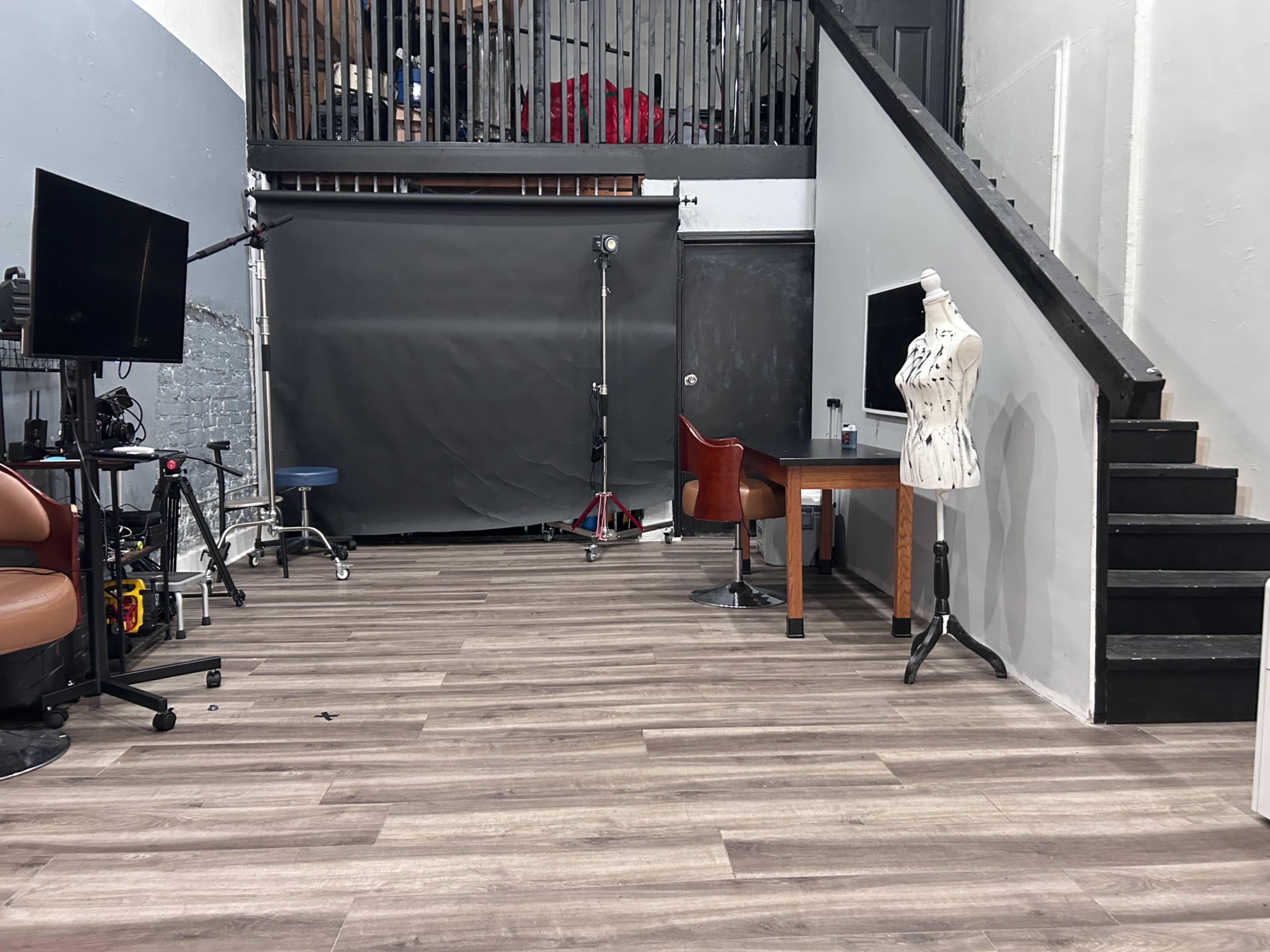 The image shows a studio space with wooden flooring, featuring a mannequin, a table, and two screens, alongside a staircase and backdrop at the rear.
