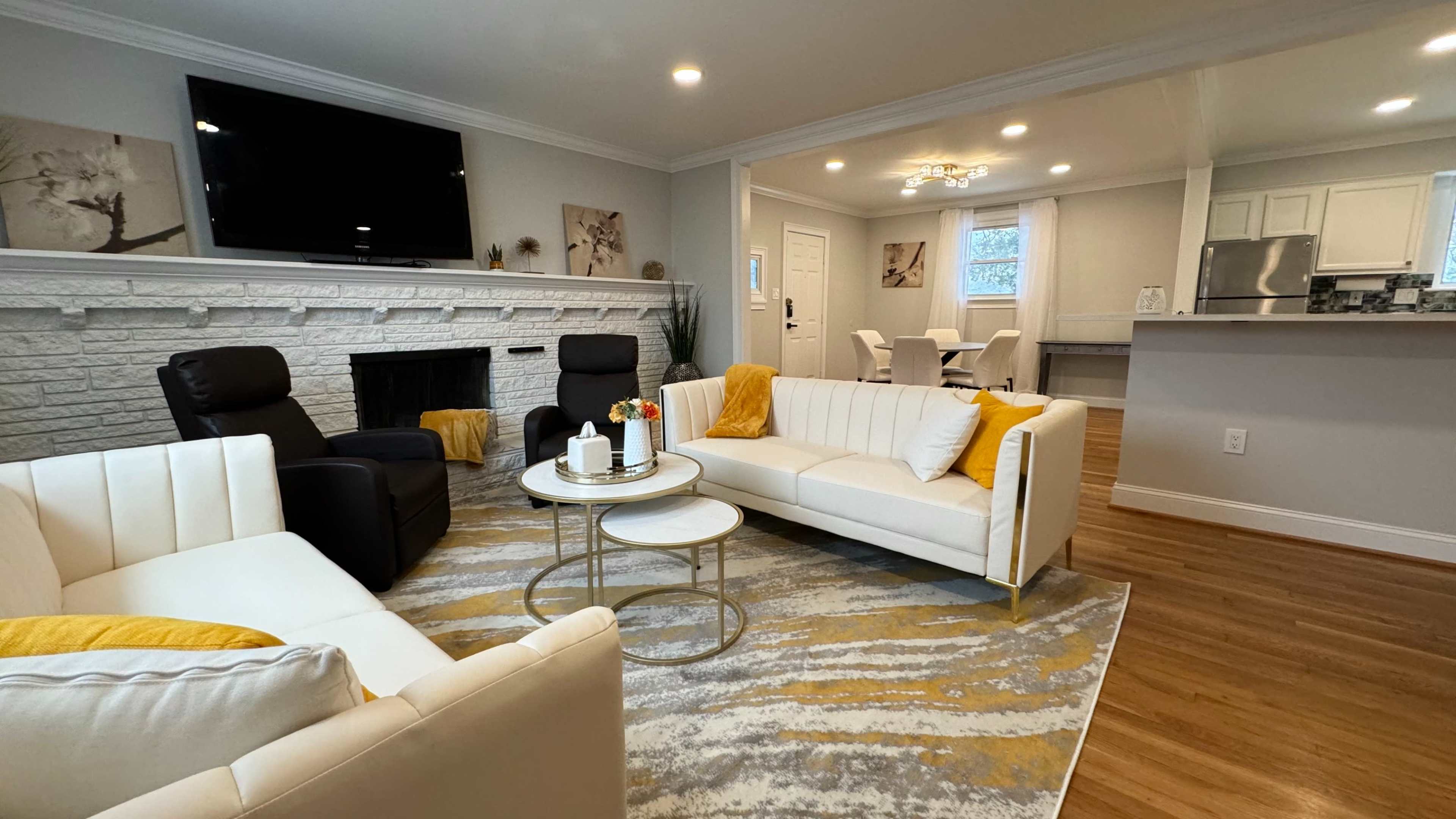 A living room with a white sofa, two armchairs, a circular coffee table, and a fireplace, all arranged around a patterned rug under overhead lighting.