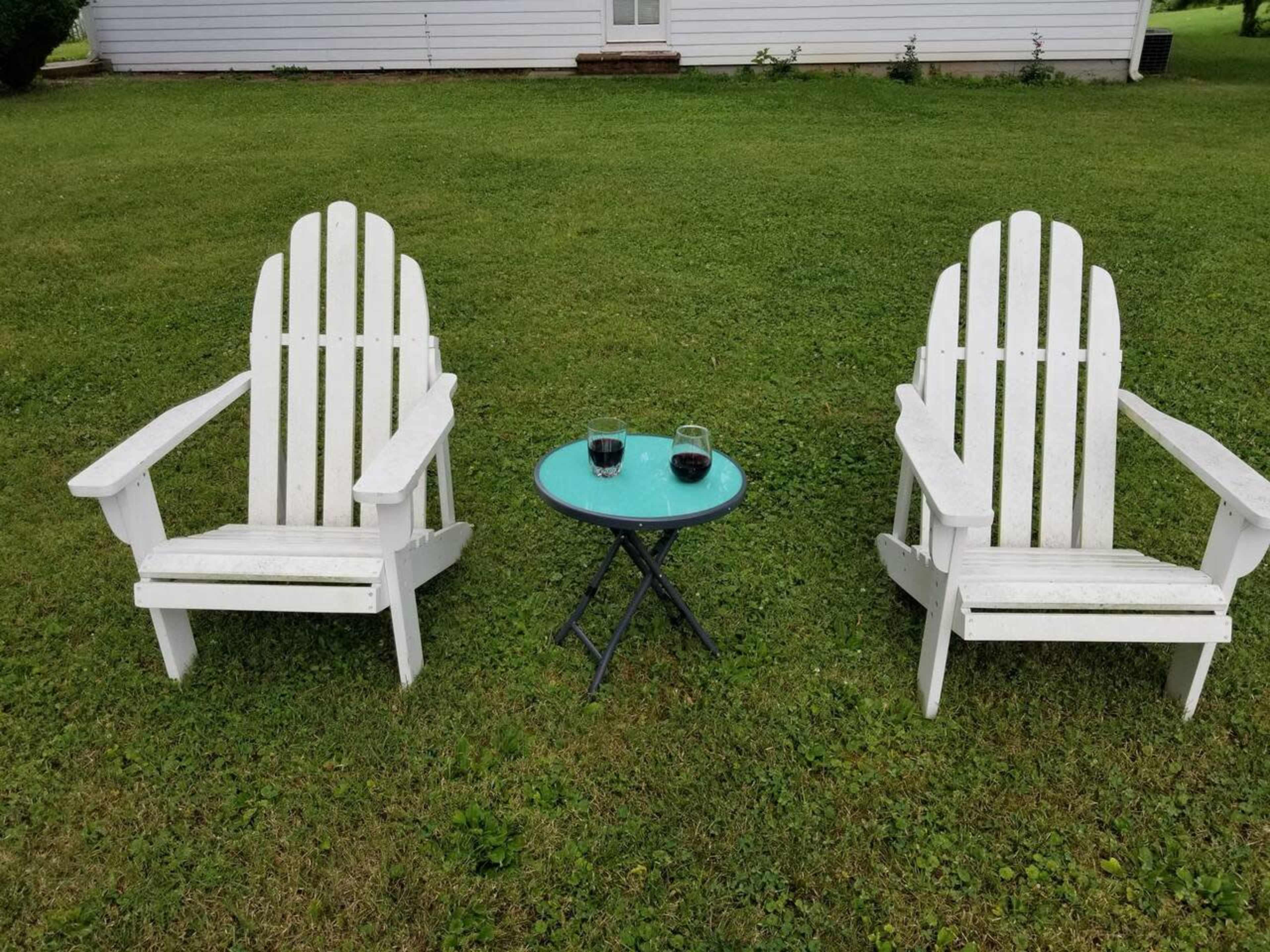 Two white Adirondack chairs are positioned on a grassy lawn beside a small, round table with two glasses on it.