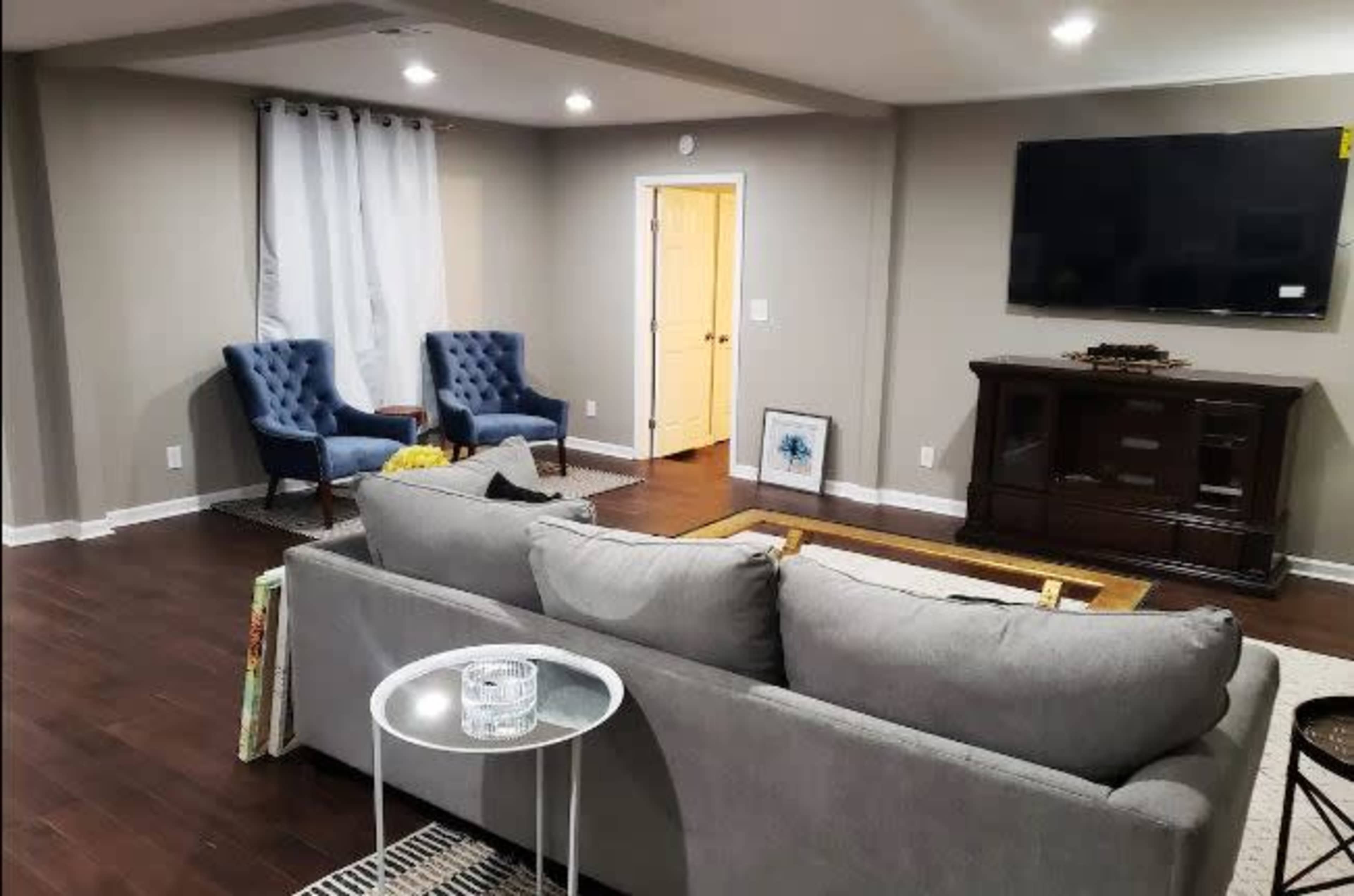 A cozy living room features a gray sofa facing a wooden entertainment unit and two blue armchairs, with a television mounted on the wall and light-colored curtains framing a nearby window.