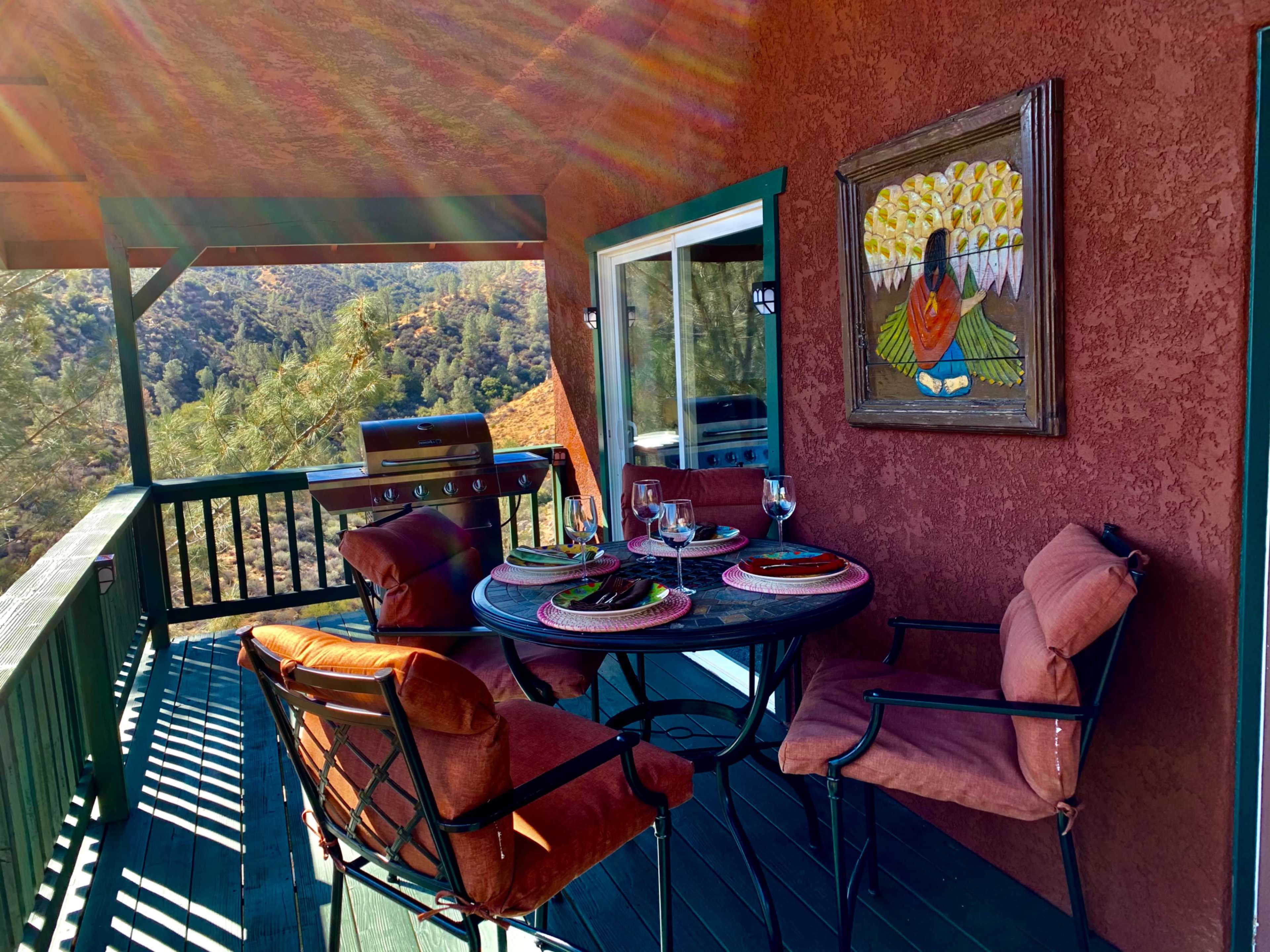A furnished balcony features a round table set for two, with a grill and a colorful painting on the wall, surrounded by trees and hills in the background.