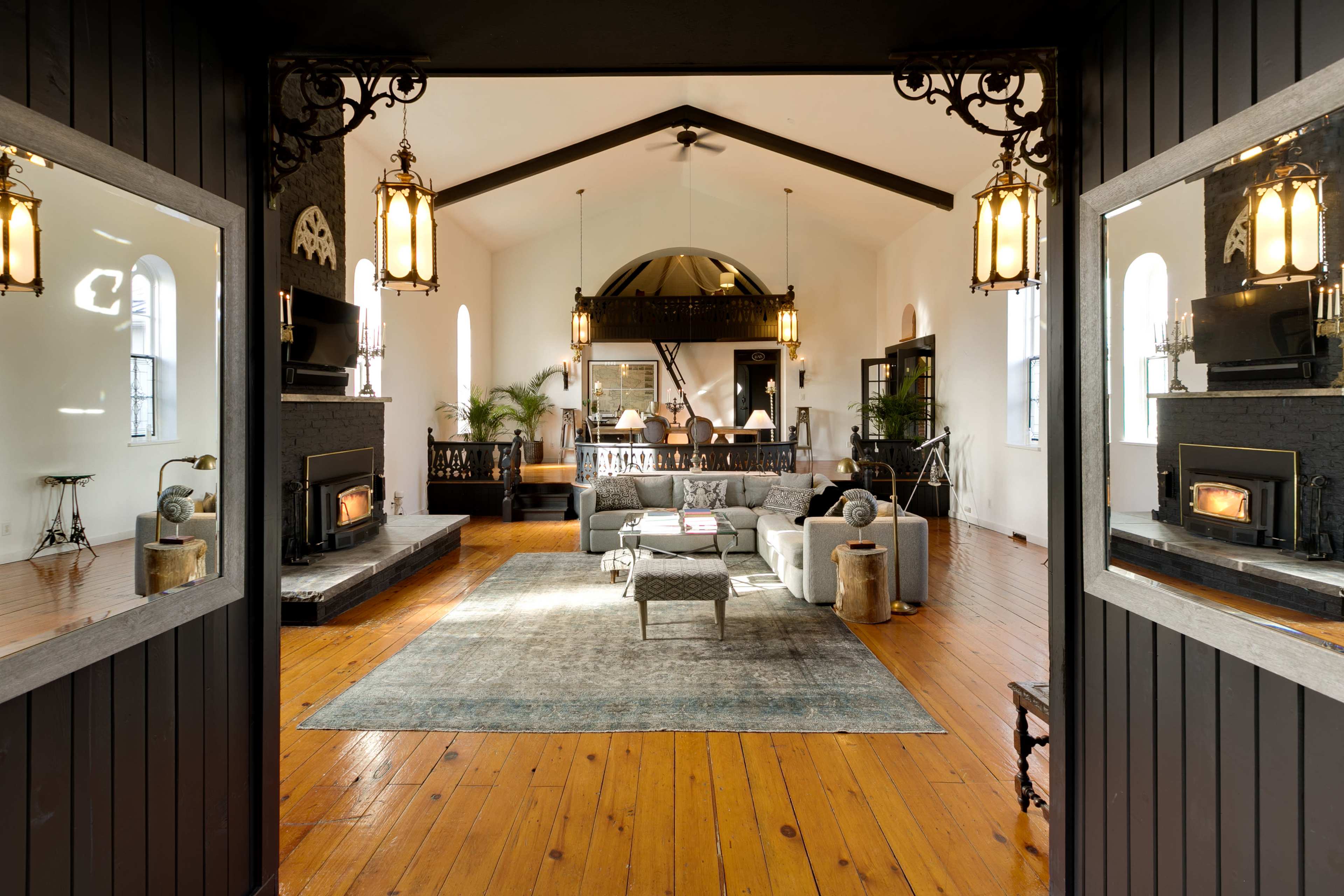 A large, open living room features a central seating area with a gray sofa and a coffee table, flanked by two decorative fireplaces and illuminated by hanging light fixtures.