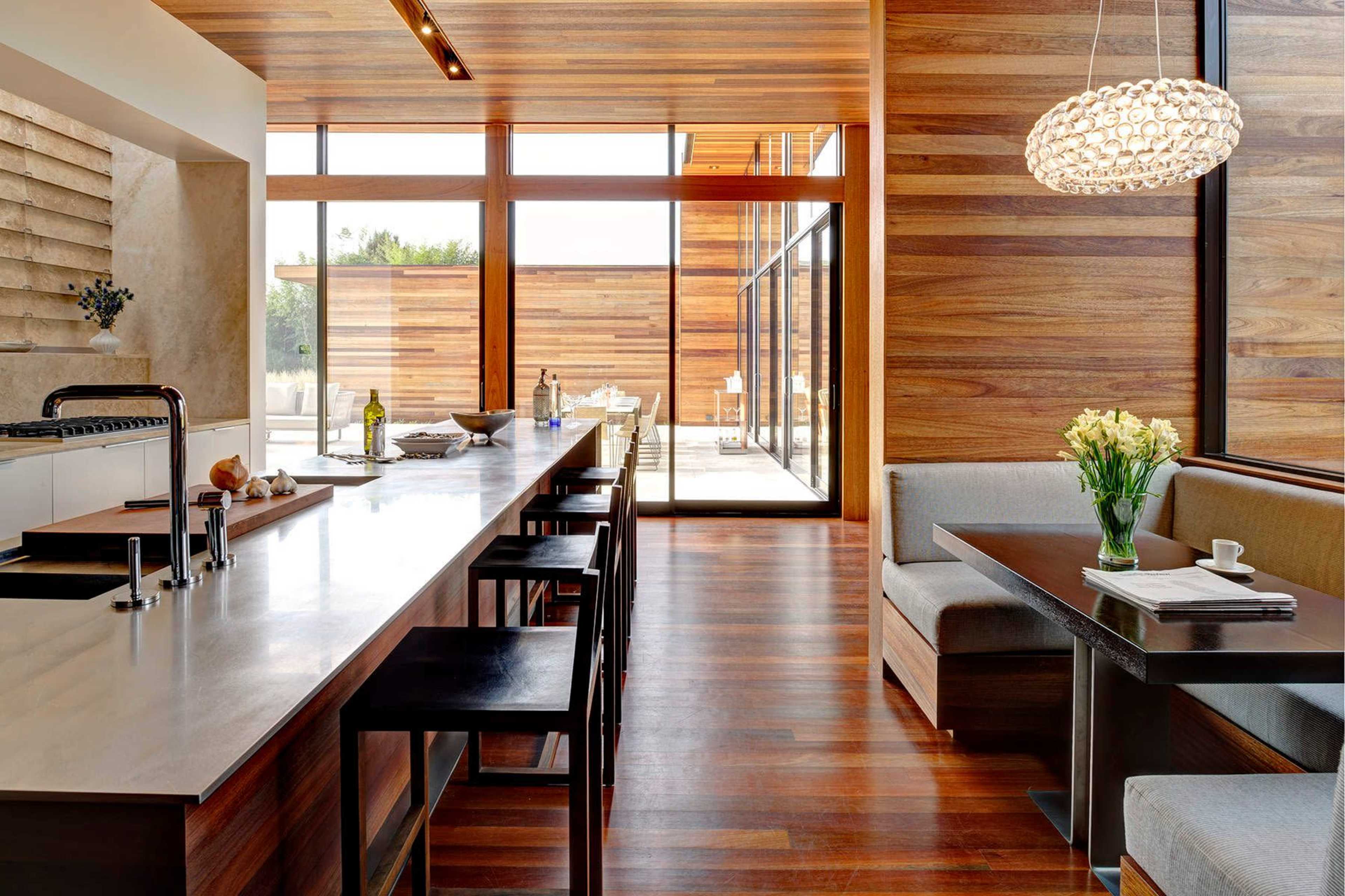 A modern kitchen with wooden walls, a long island counter, and a seating area beside large windows.