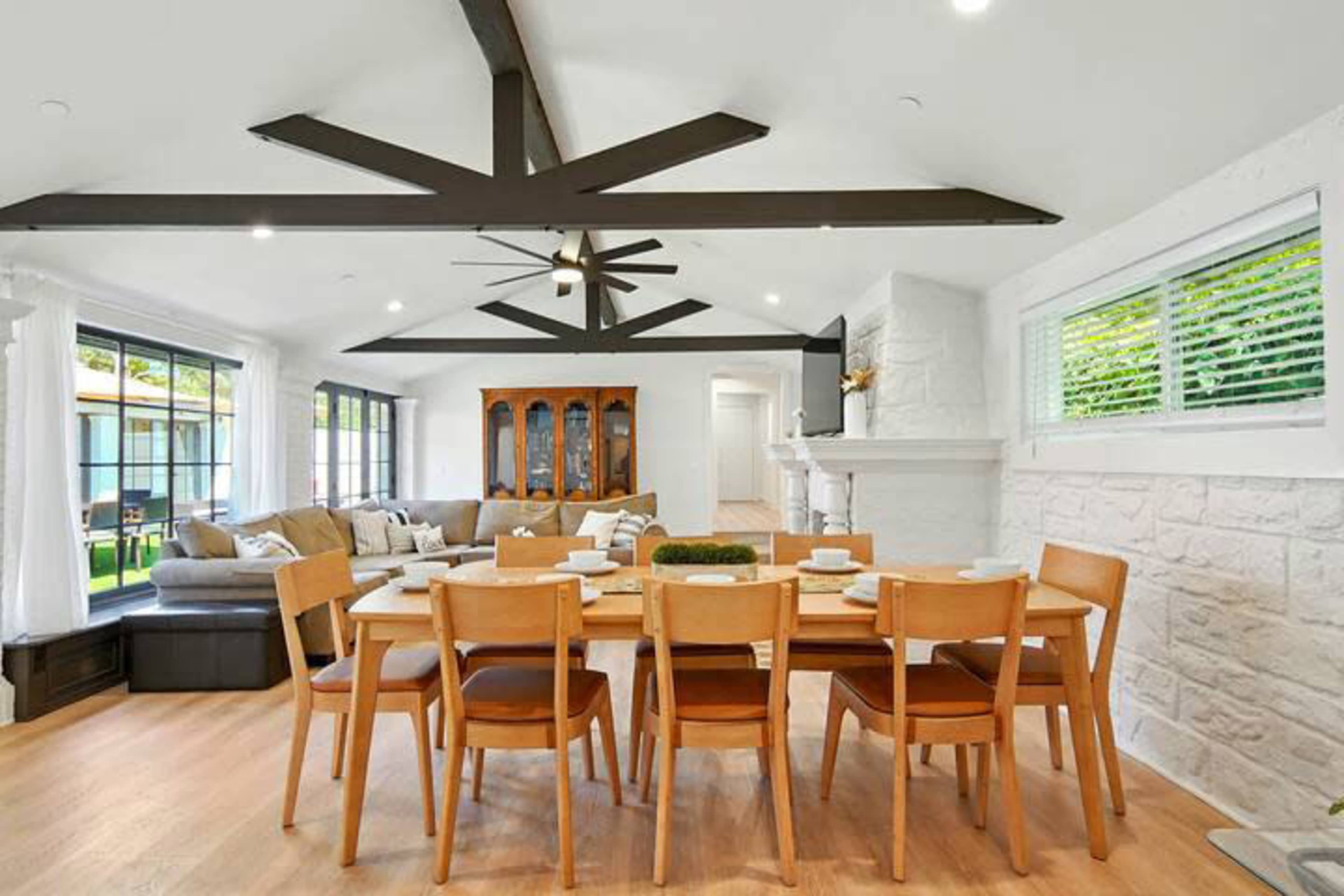 A spacious dining area features a wooden table surrounded by eight chairs, with a living room and large windows in the background.