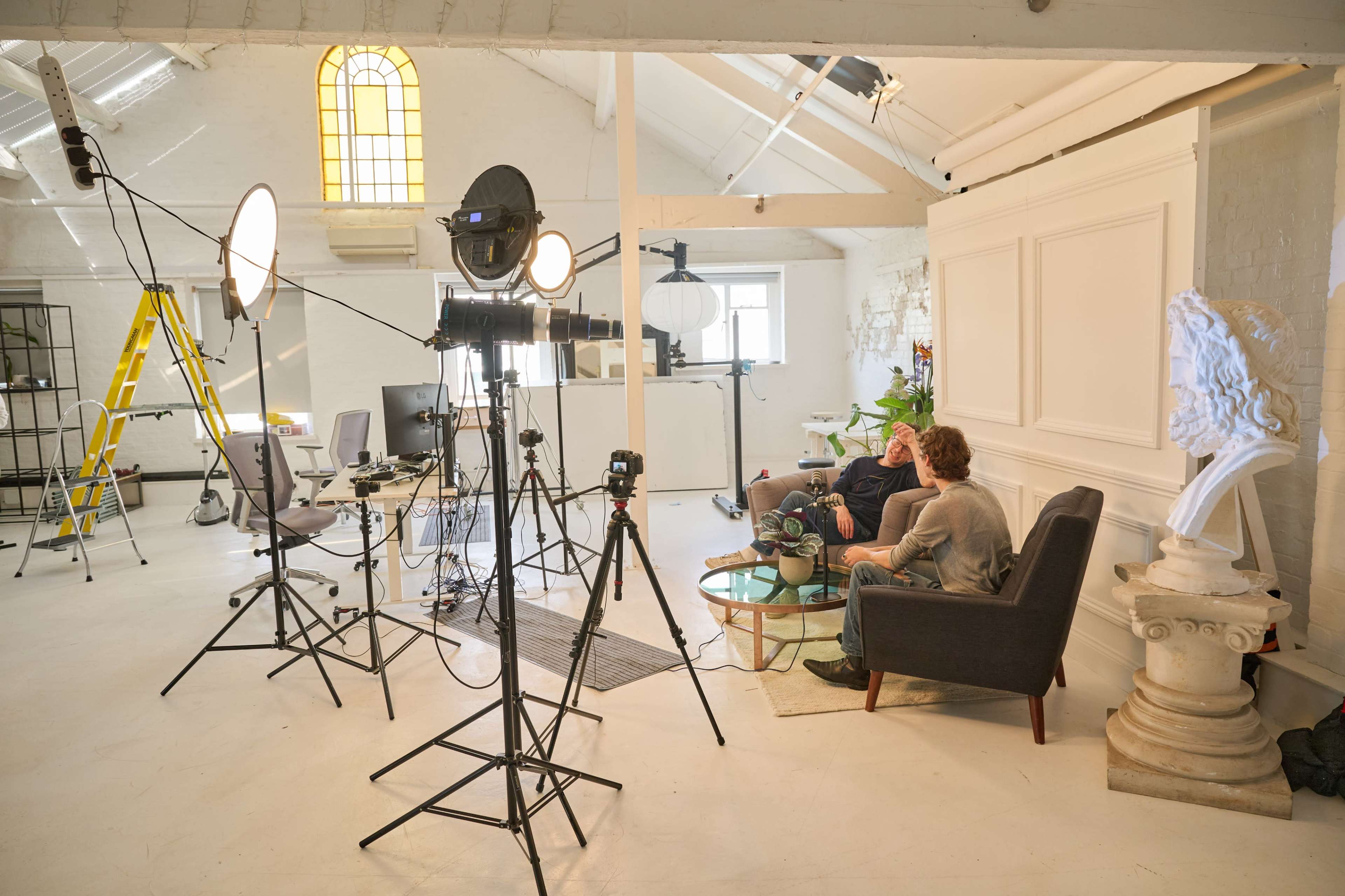 A well-lit studio space features cameras, lighting equipment, a ladder, and two individuals seated on a couch engaged in conversation.