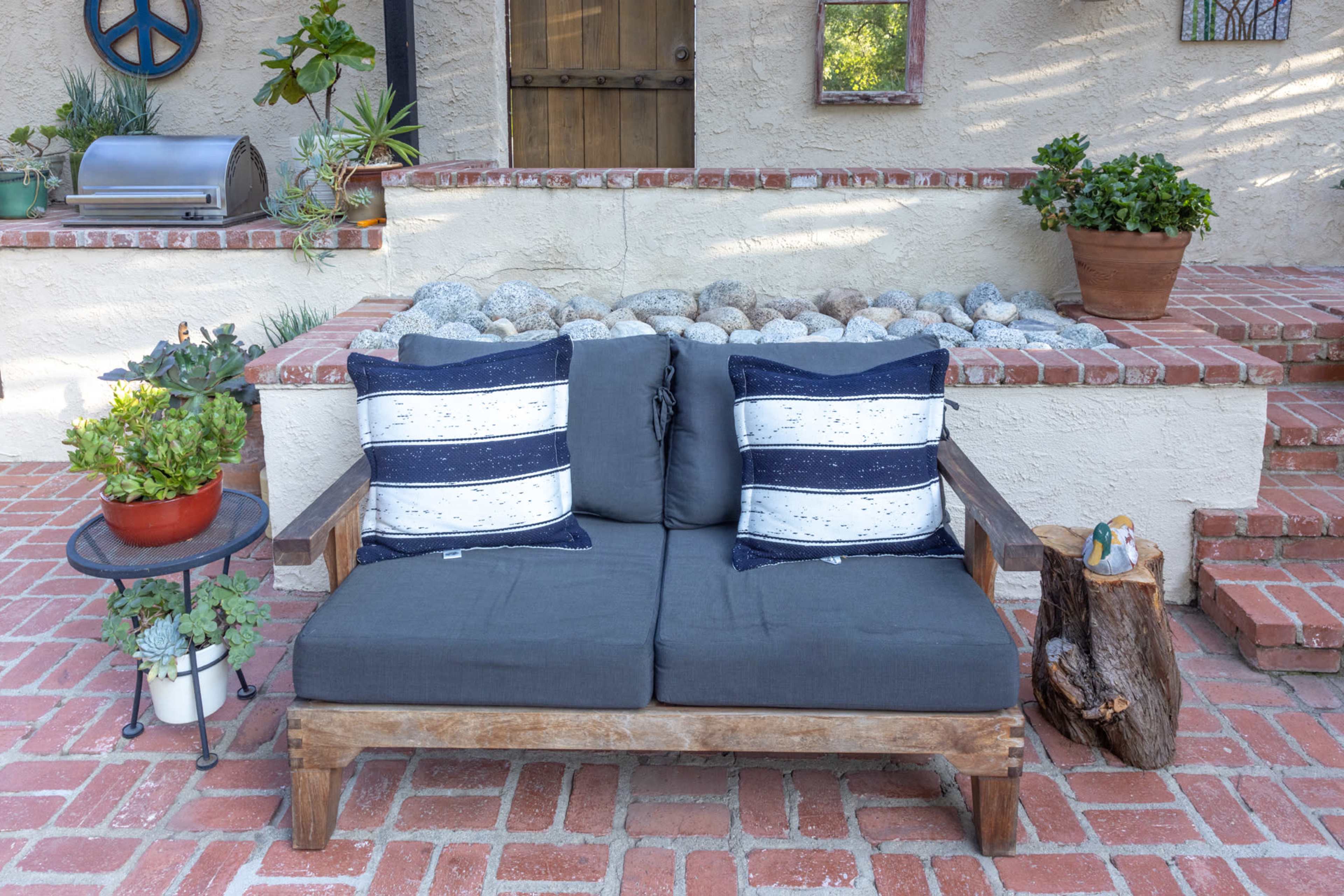 A wooden loveseat with gray cushions and decorative pillows is positioned in front of a brick wall adorned with potted plants and a small table.
