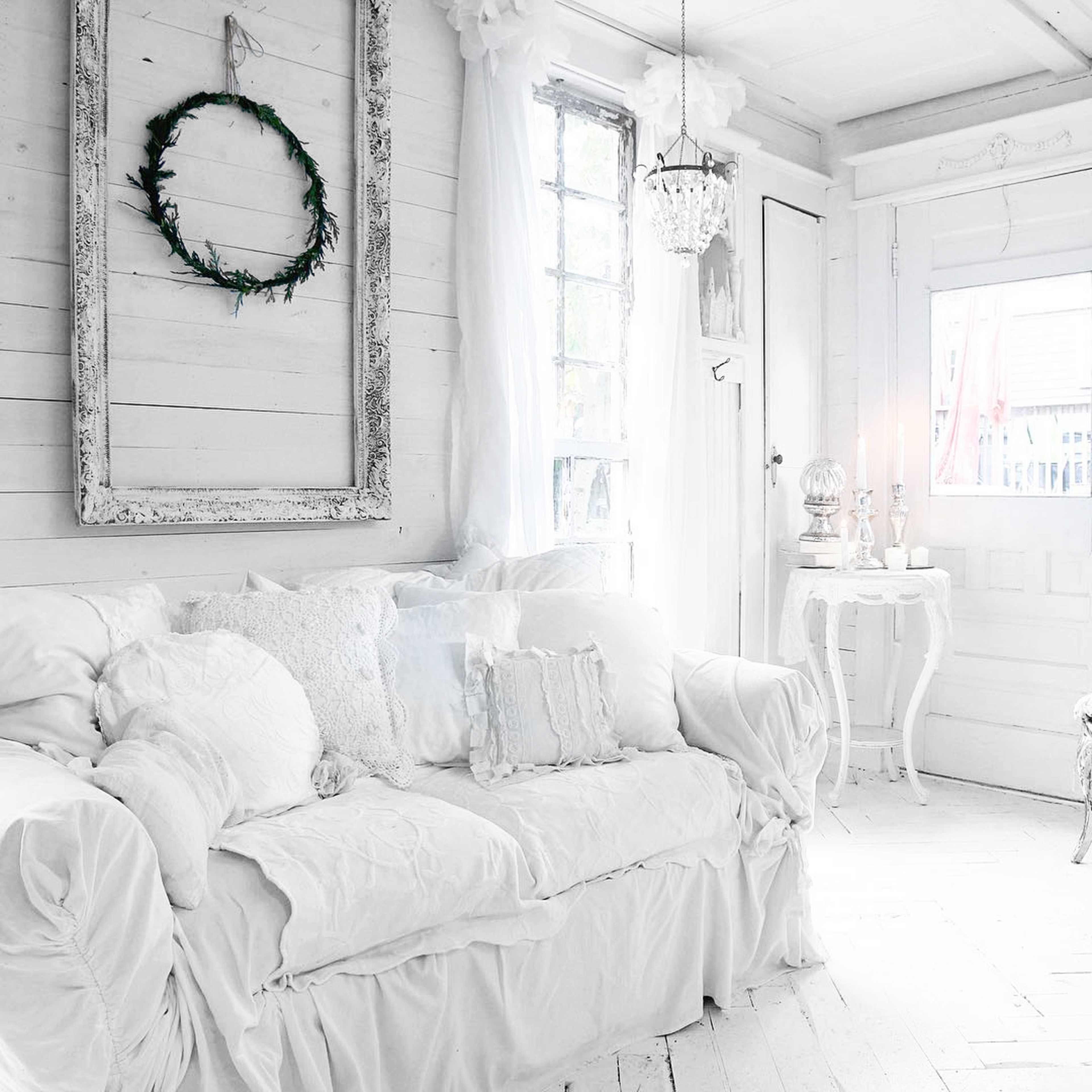 A light-filled room with white walls, a white sofa covered in soft cushions, and a decorative wreath hung on the wall.
