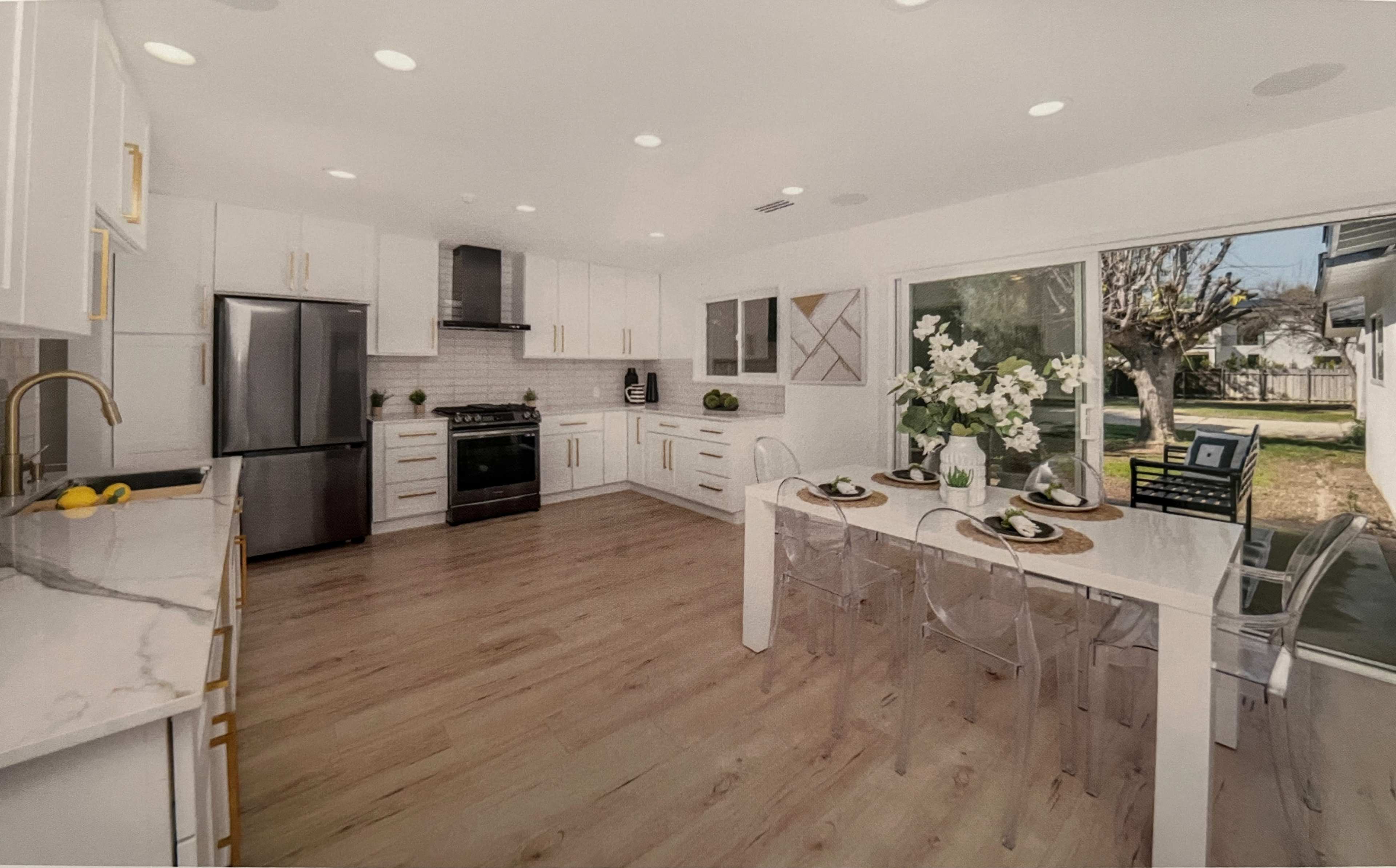 A modern kitchen with white cabinets, stainless steel appliances, and a dining area featuring a clear table and chairs, illuminated by natural light from a large window.