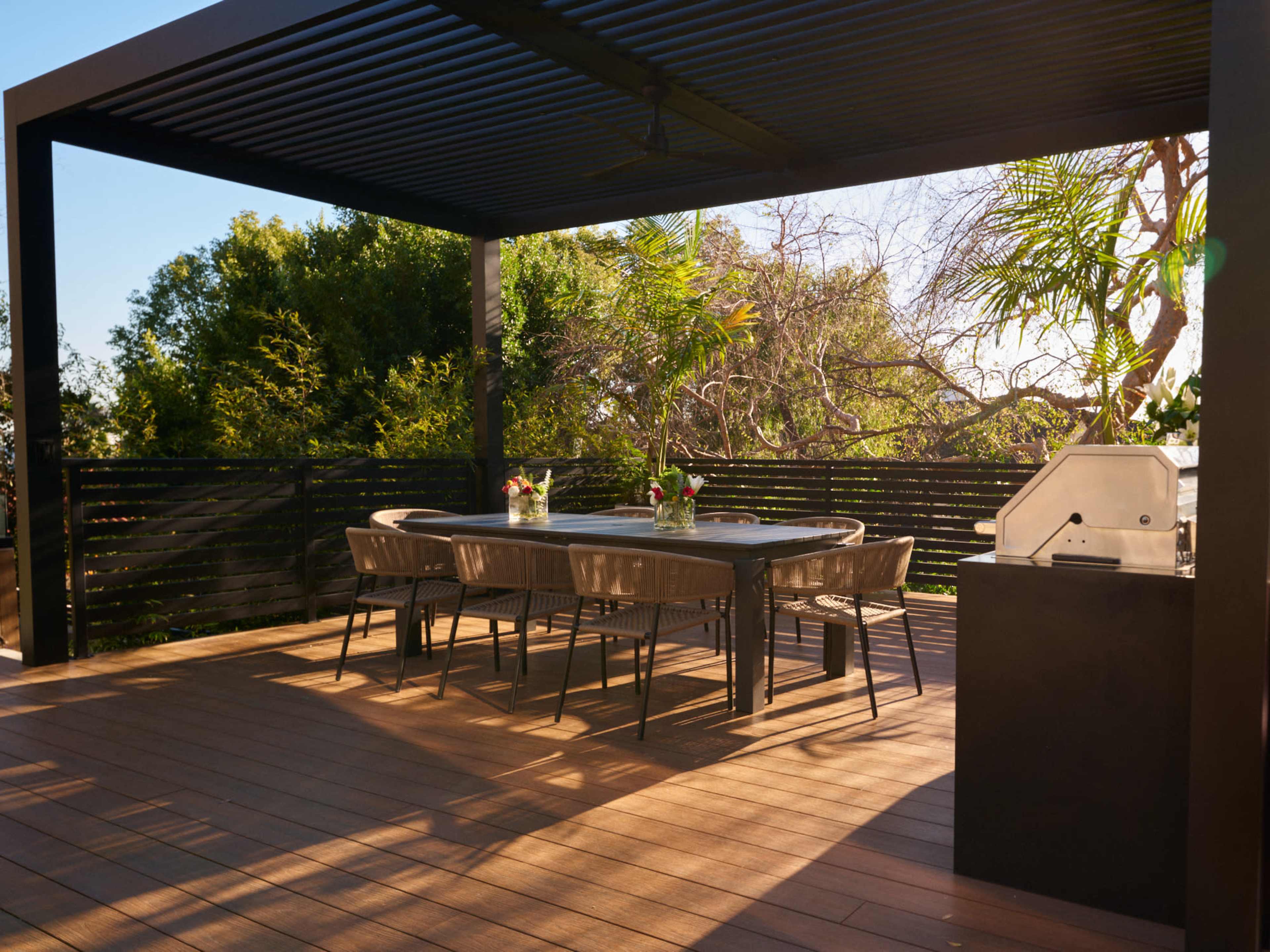 A modern outdoor dining area features a table and chairs under a pergola, with a barbecue grill nearby and surrounded by greenery.
