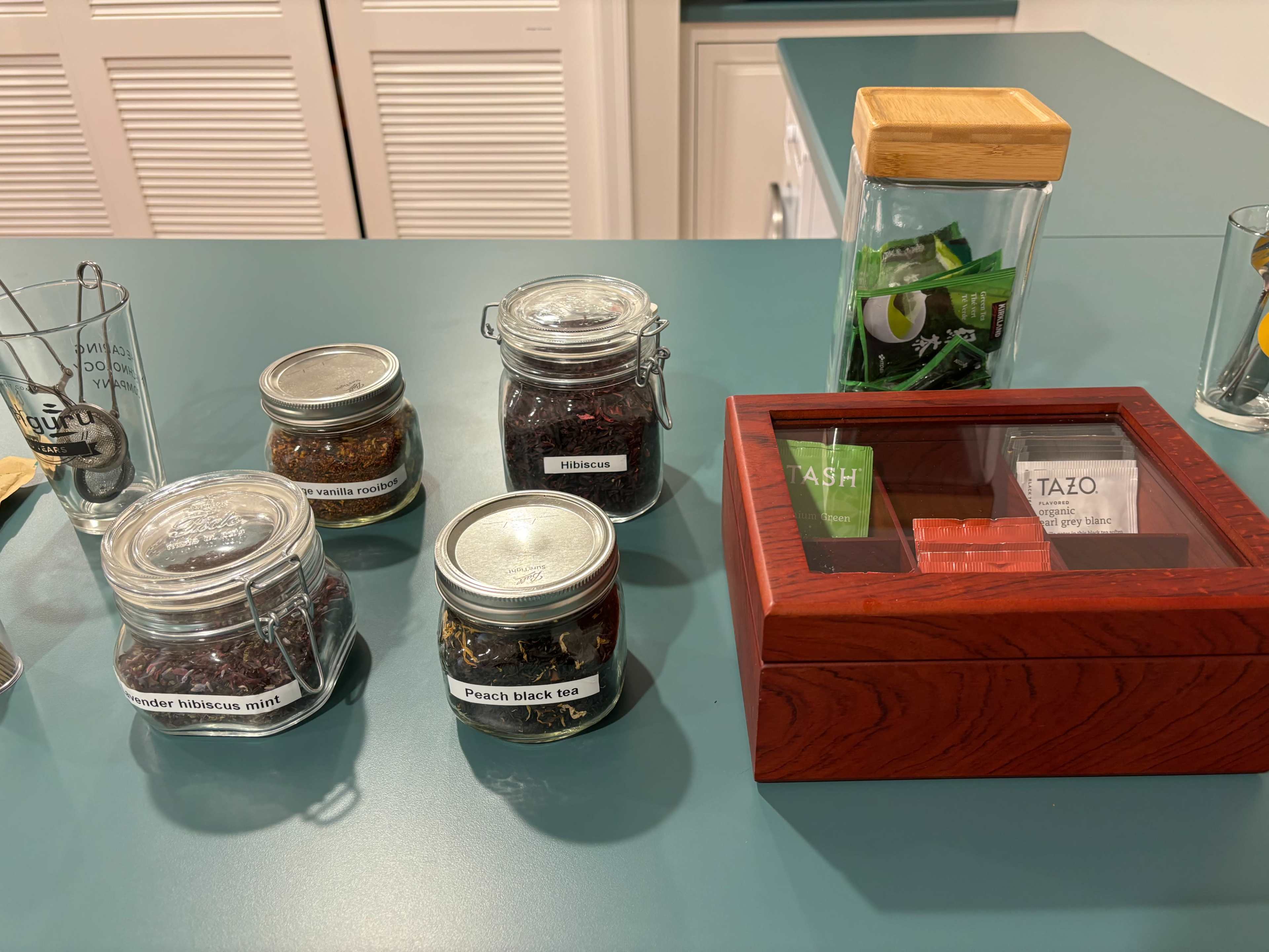 A countertop with several labeled jars of loose tea and a wooden box containing various tea bags.