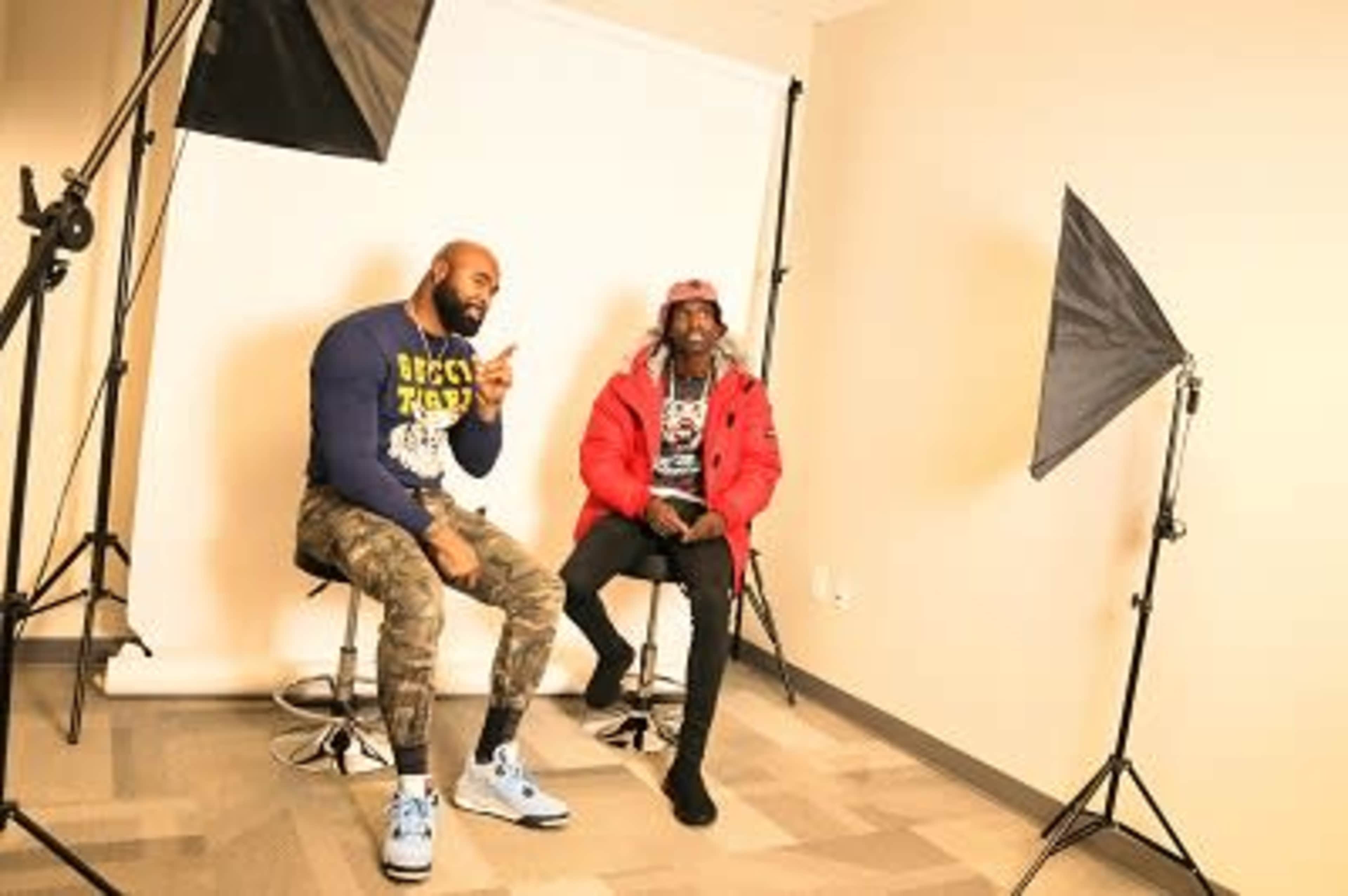 Two men sit on stools in a photography studio, surrounded by lighting equipment and a neutral backdrop.