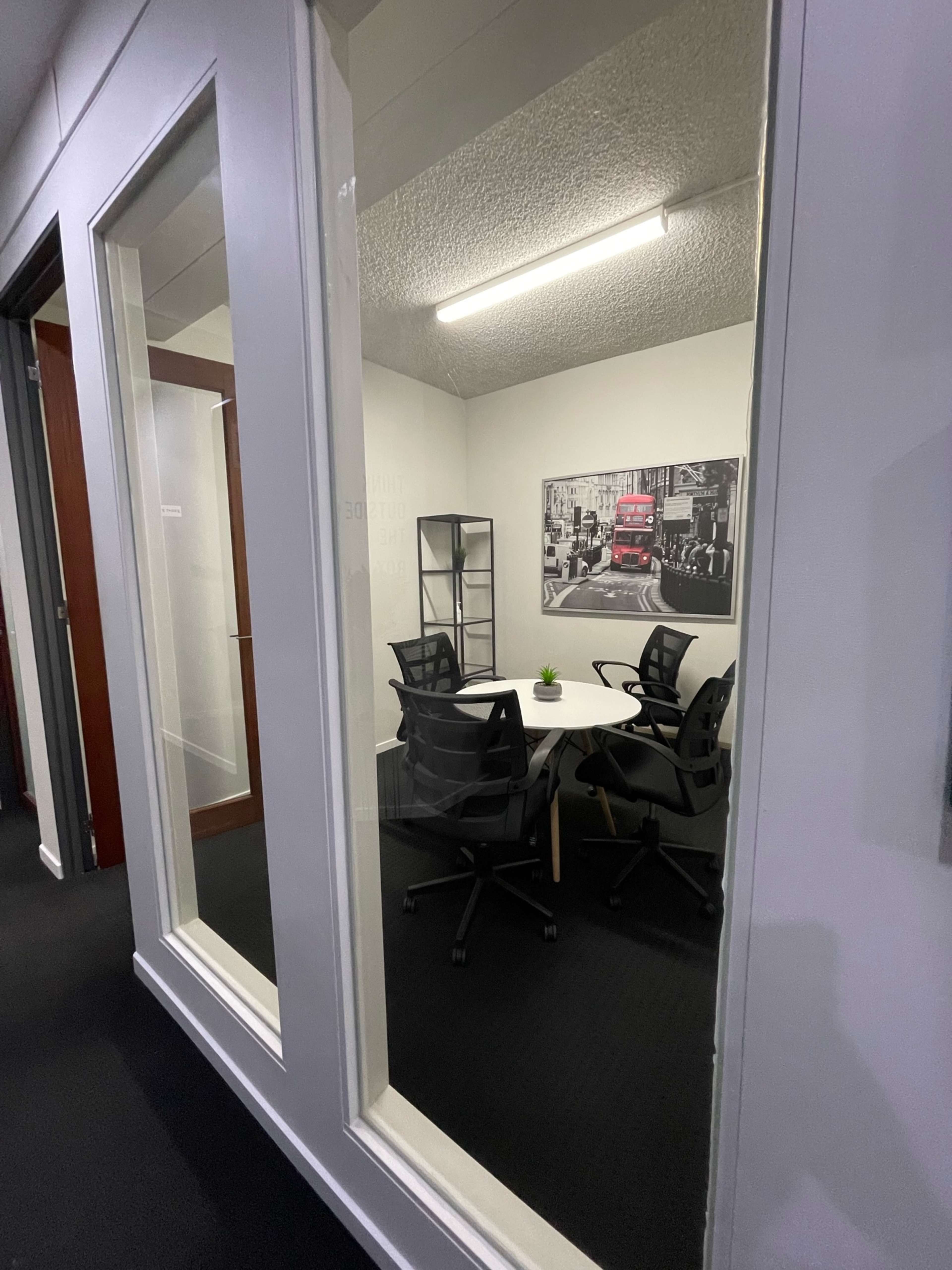 The image shows a modern conference room with a round table and four black chairs, visible through large glass windows.
