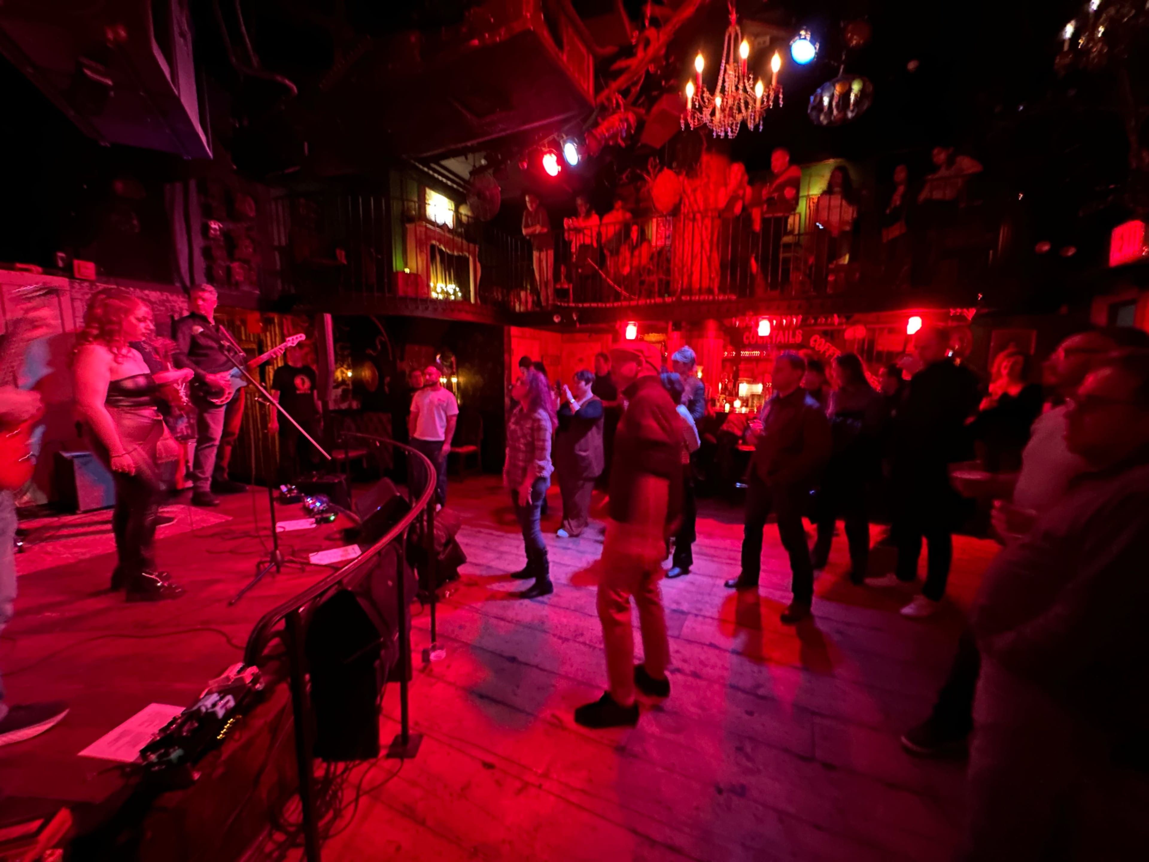 A band performs on stage while people dance and socialize in a vibrant, dimly lit venue decorated with red lighting.